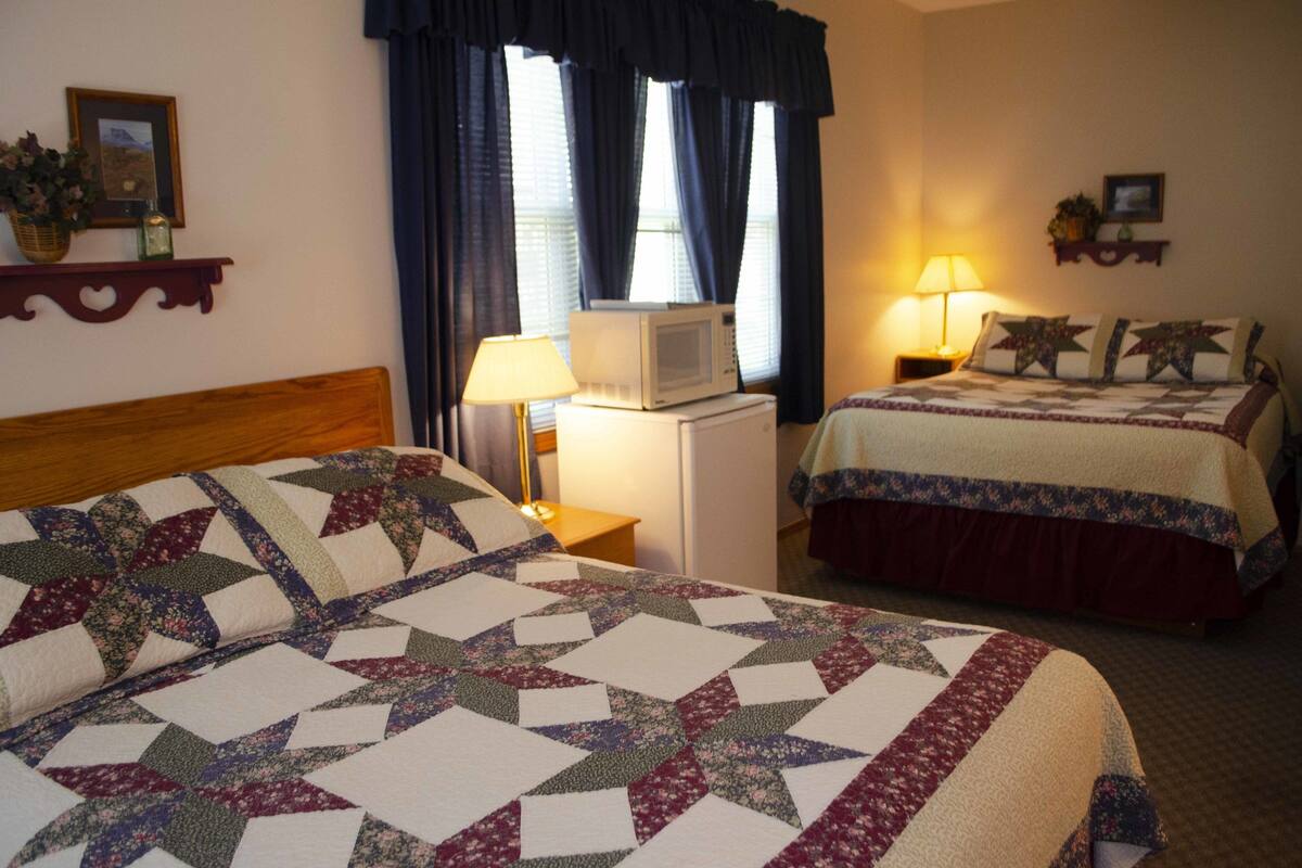 A hotel room with two beds, a microwave, a nightstand with a lamp, and a window with curtains.