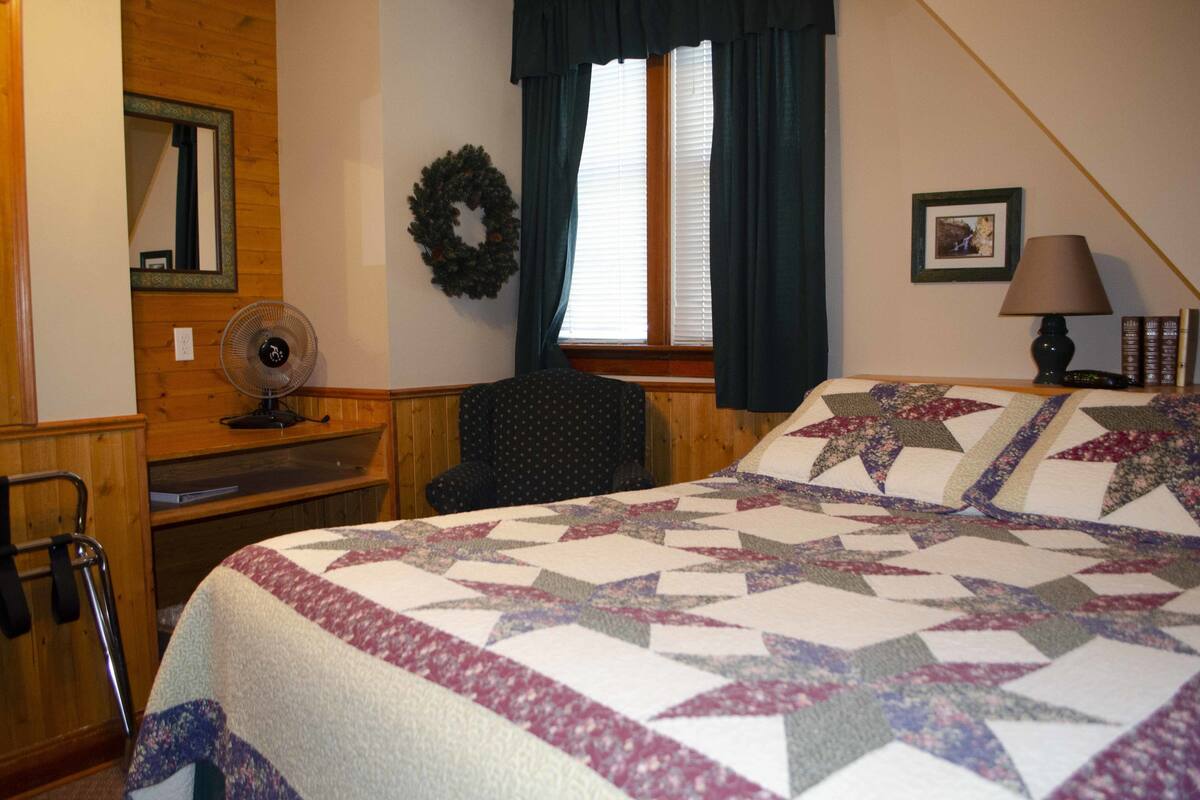 A bedroom with a bed, a chair, a fan, a wheelchair, a window with curtains, a wall-mounted mirror, a picture frame, and a lamp.