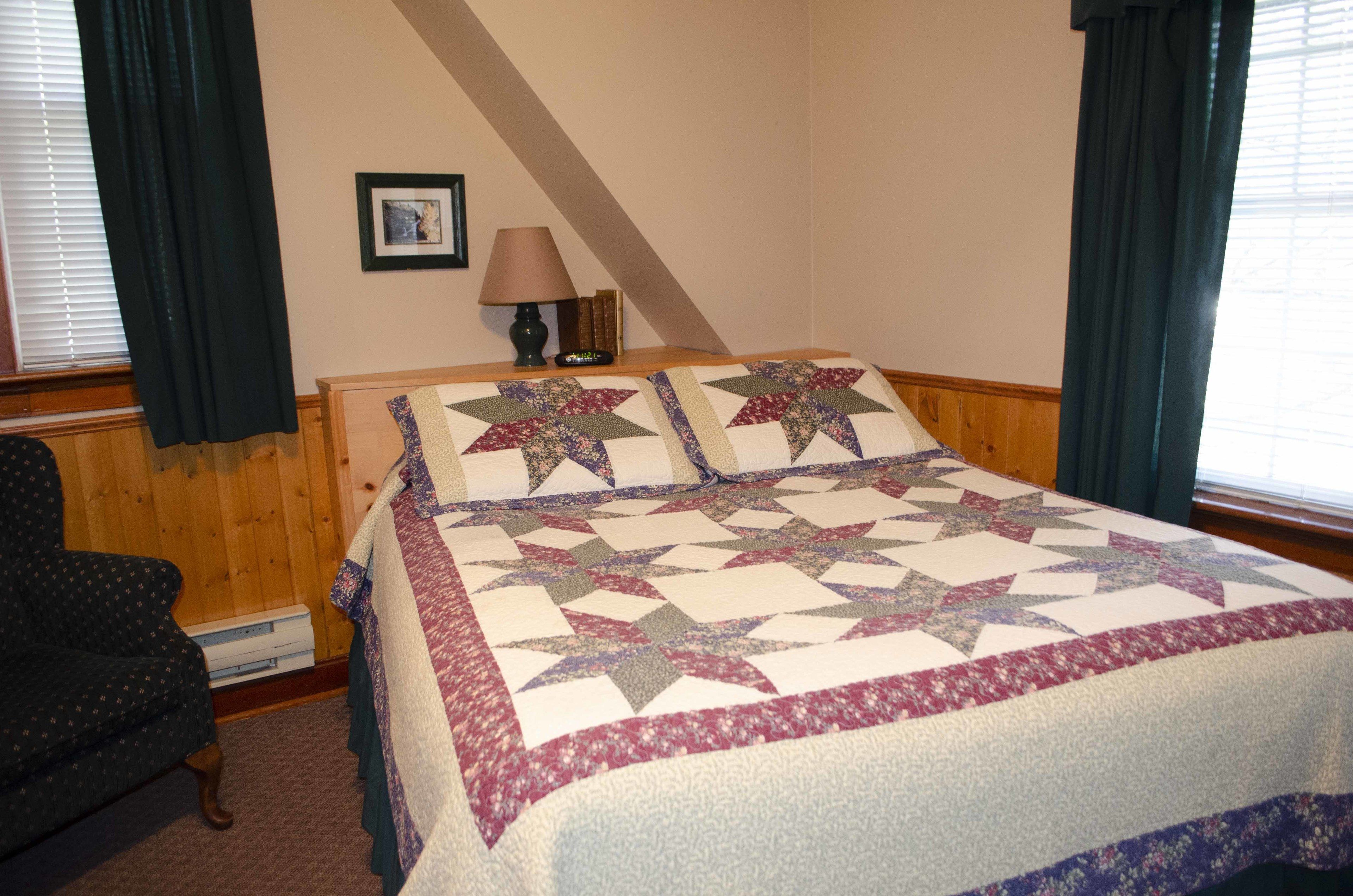 A bedroom with a large bed, a wooden headboard, a nightstand with a lamp, a framed picture on the wall, and a window with curtains.