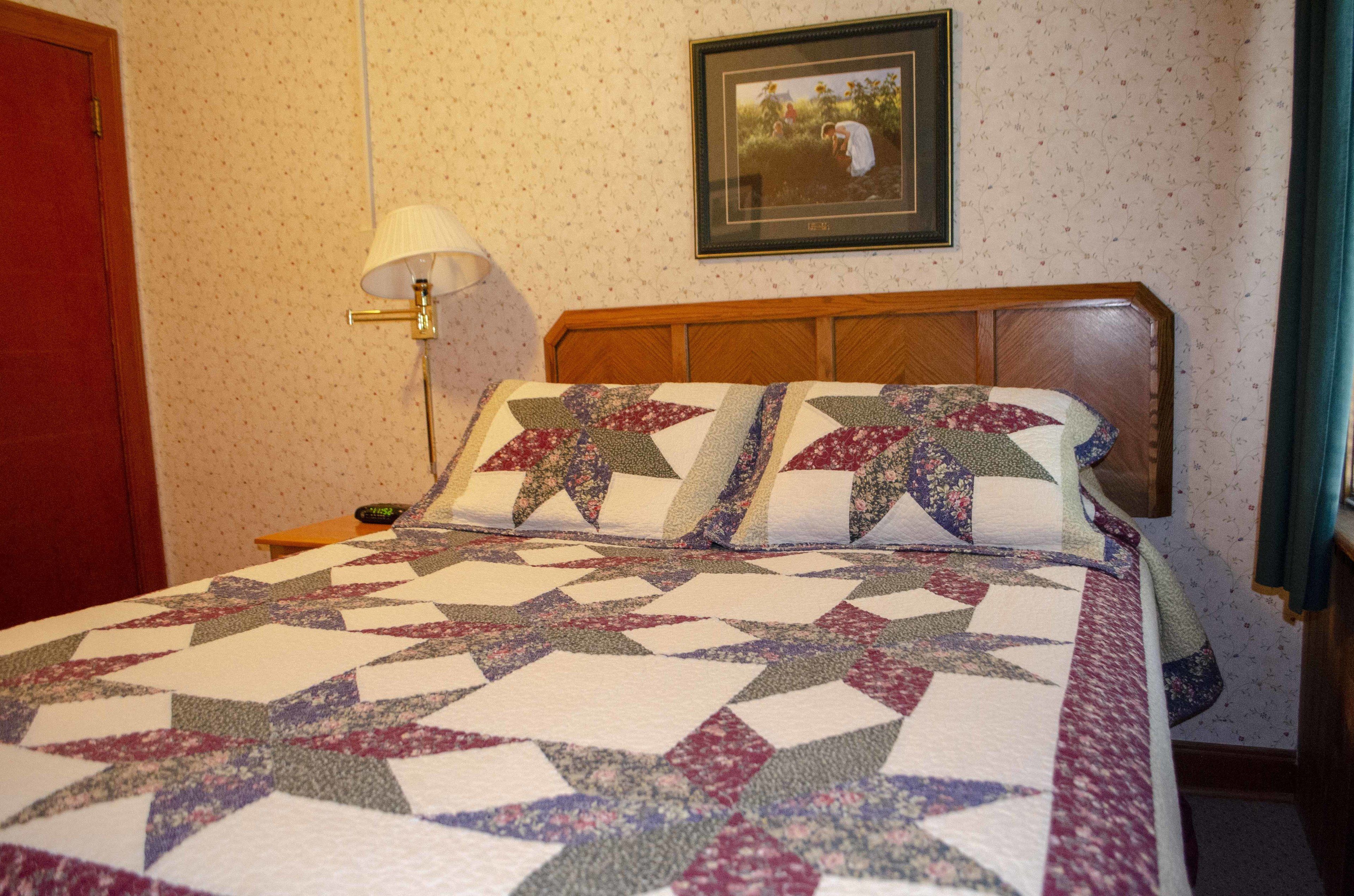 A neatly made bed with a patterned quilt, a bedside lamp, and a framed picture on the wall.