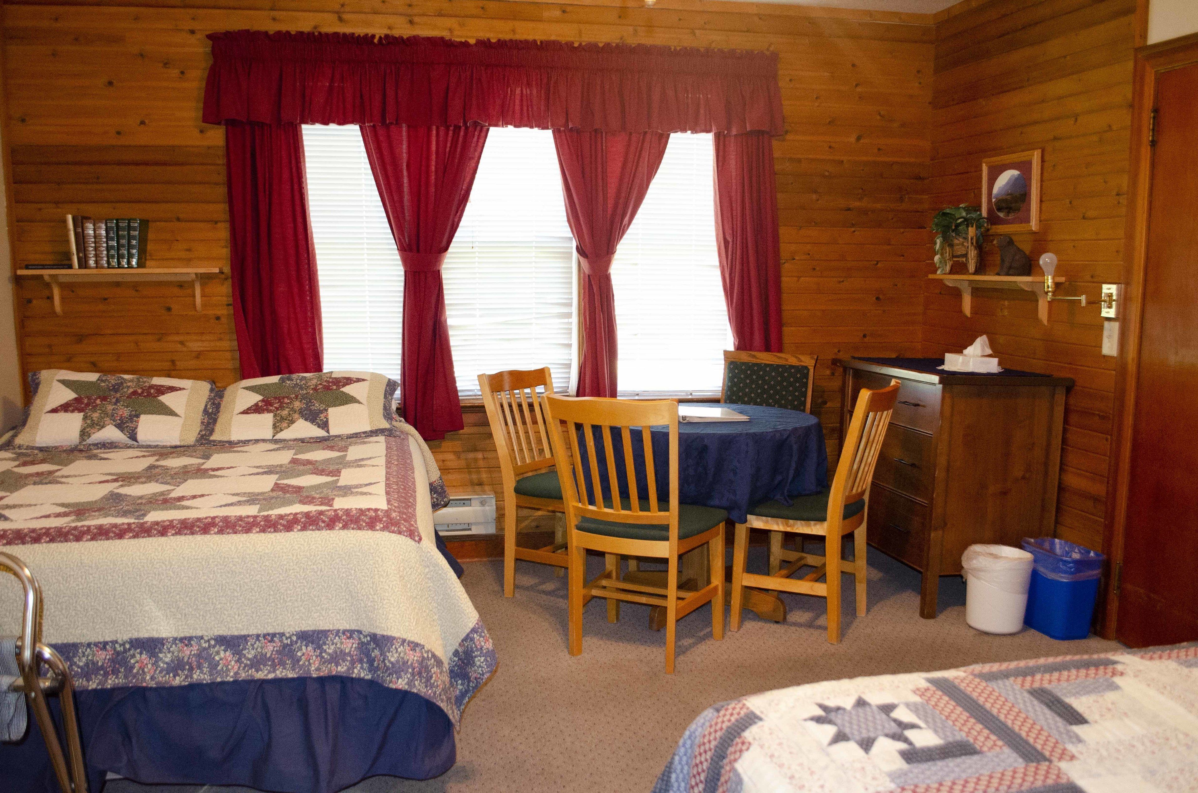 A cozy cabin room with two beds, a dining table, chairs, a small desk, and a window with curtains.