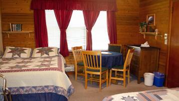 A cozy cabin room with two beds, a dining table, chairs, a small desk, and a window with curtains.