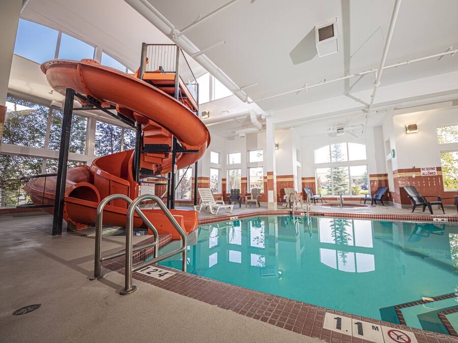 Ein Hallenbad mit Wasserrutsche, Liegestühlen und Blick auf Bäume durch große Fenster.