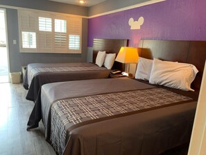 A hotel room with two beds, a purple accent wall, and a window with blinds.