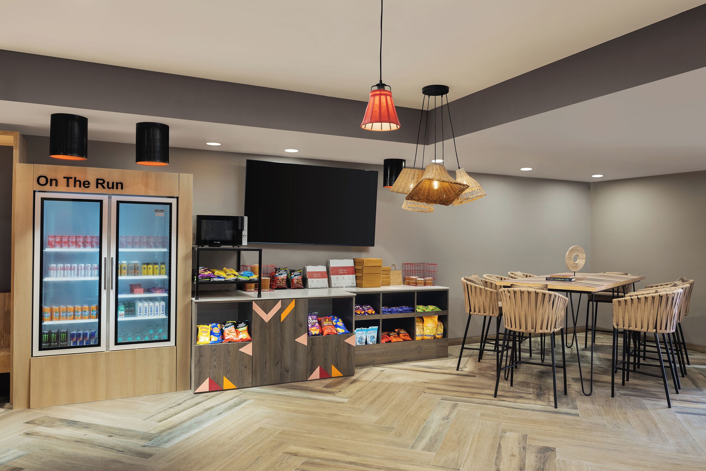 Un área de comedor moderna con una mesa y sillas de madera, un refrigerador identificado como  “On The Run” y diversos aperitivos en estantes.