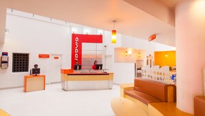 Un área de recepción con un mostrador central, dos recepcionistas, una zona de estar con un sofá y una mesa, y una pared con un gran cartel que dice 'ginger'.