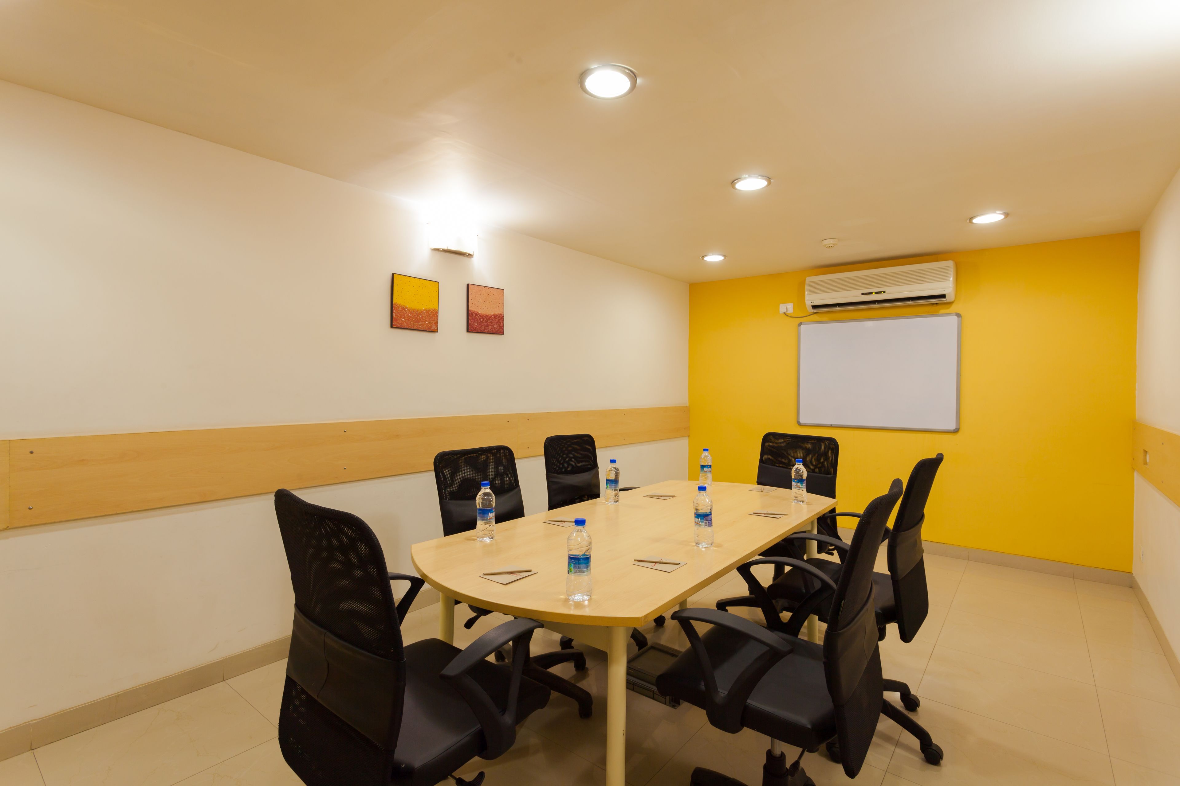 A conference room with a wooden table, black chairs, and water bottles. There is a whiteboard on the wall and an air conditioning unit.
