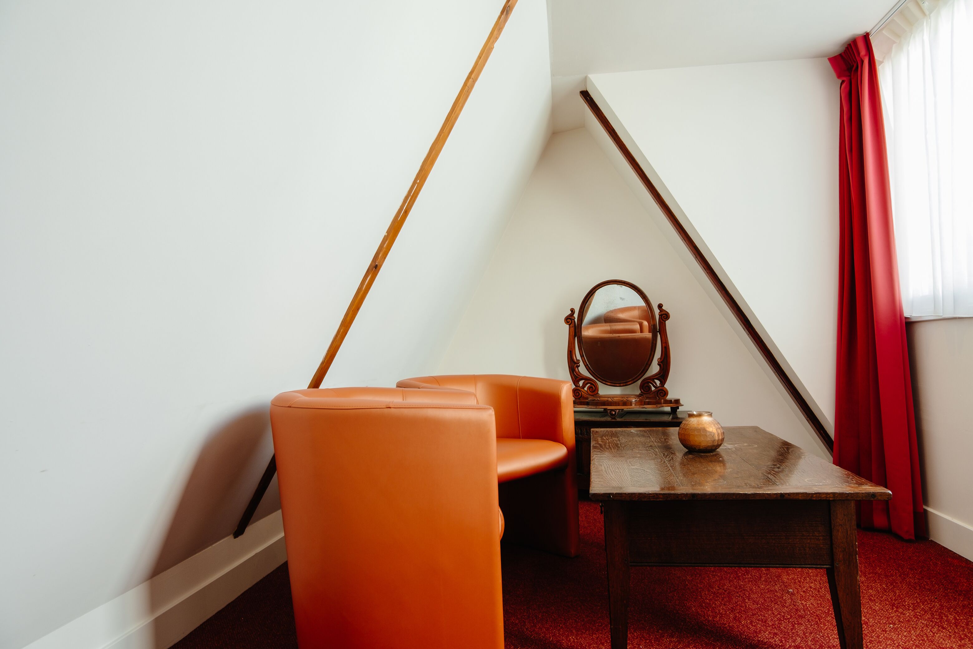 Une pièce avec un plafond en pente, une table en bois sur laquelle est posé un vase, une chaise orange et un miroir.
