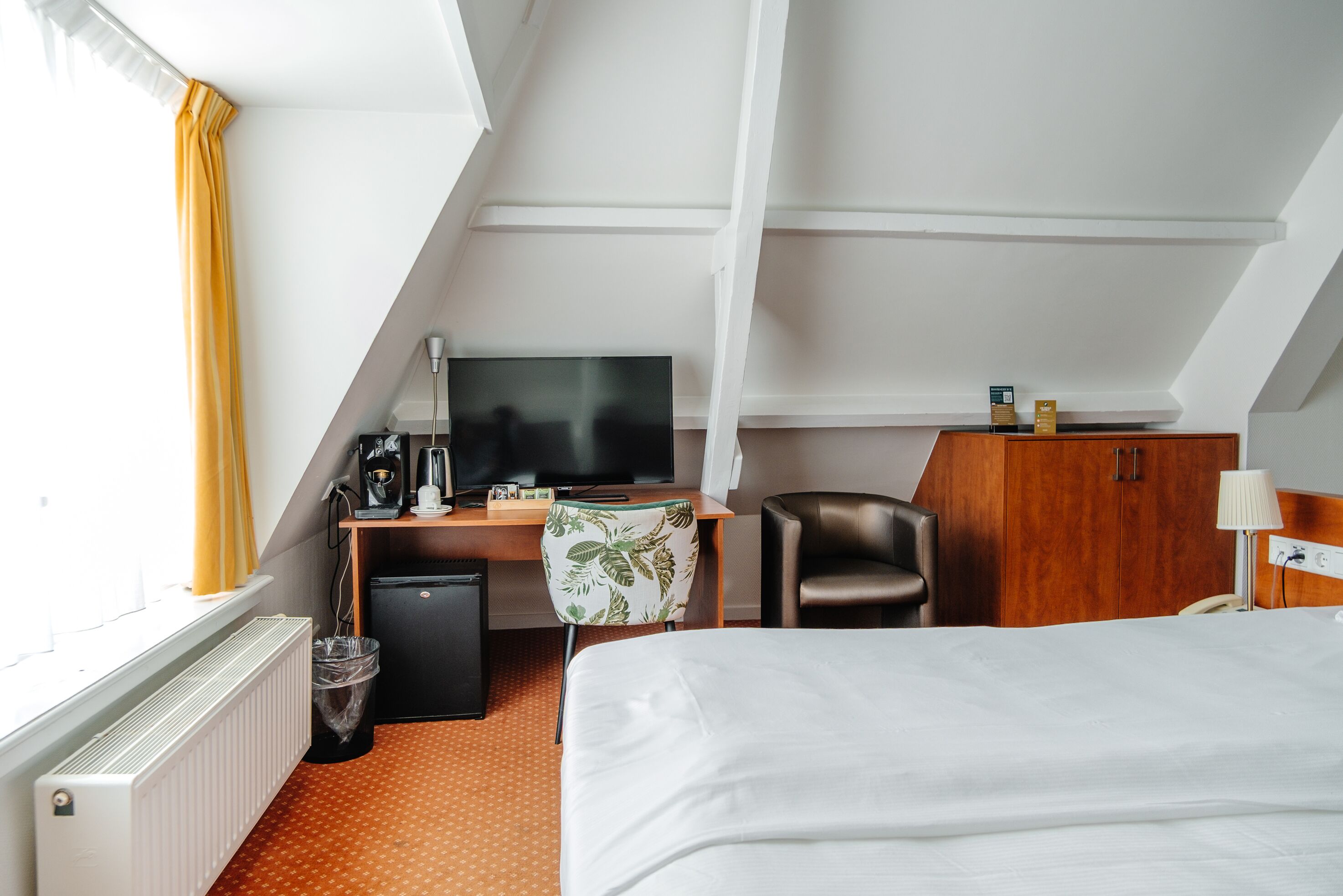 A hotel room with a bed, a desk with a TV, a chair, a wardrobe, and a radiator.