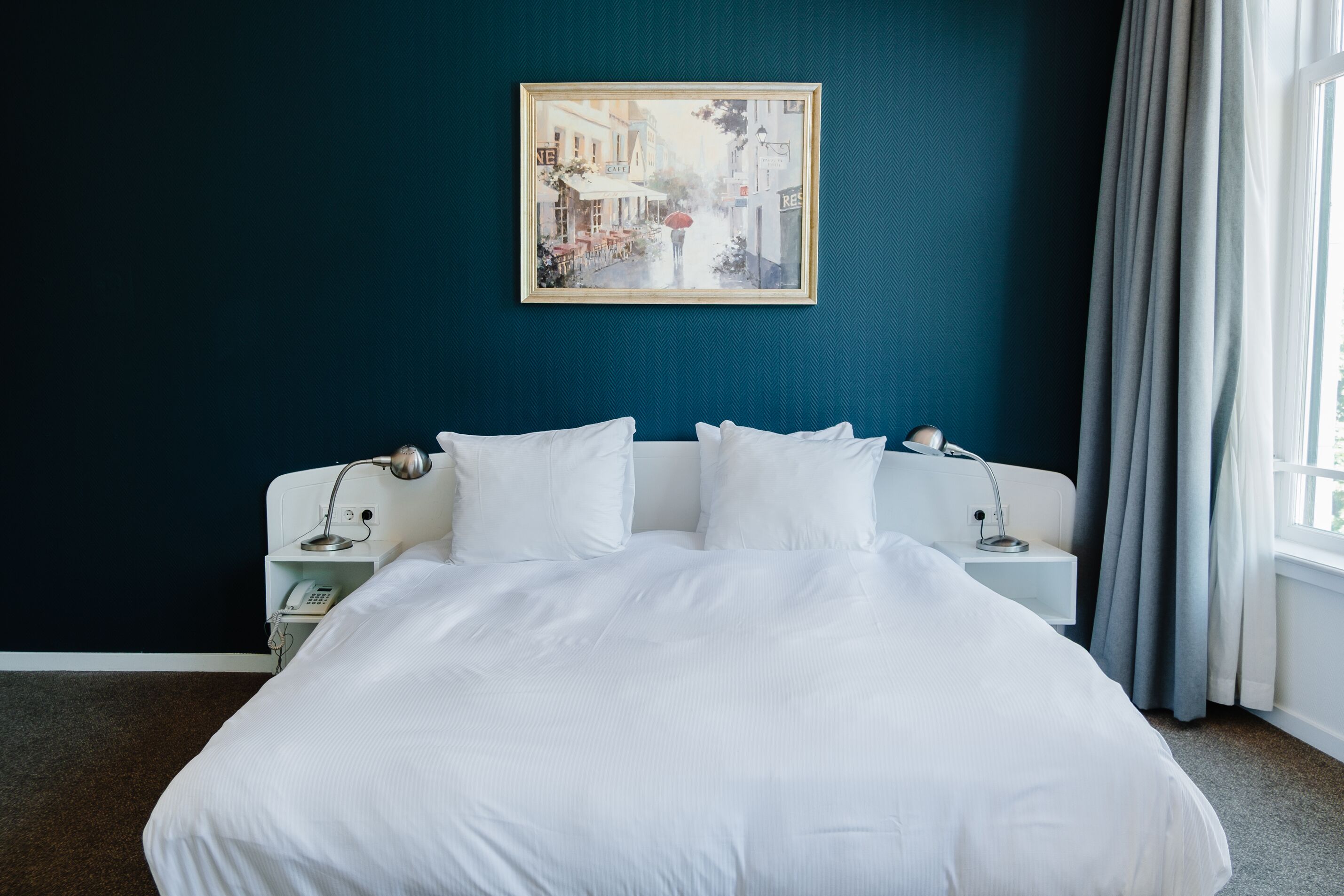 A neatly made bed with white bedding in a room with a dark teal wall.