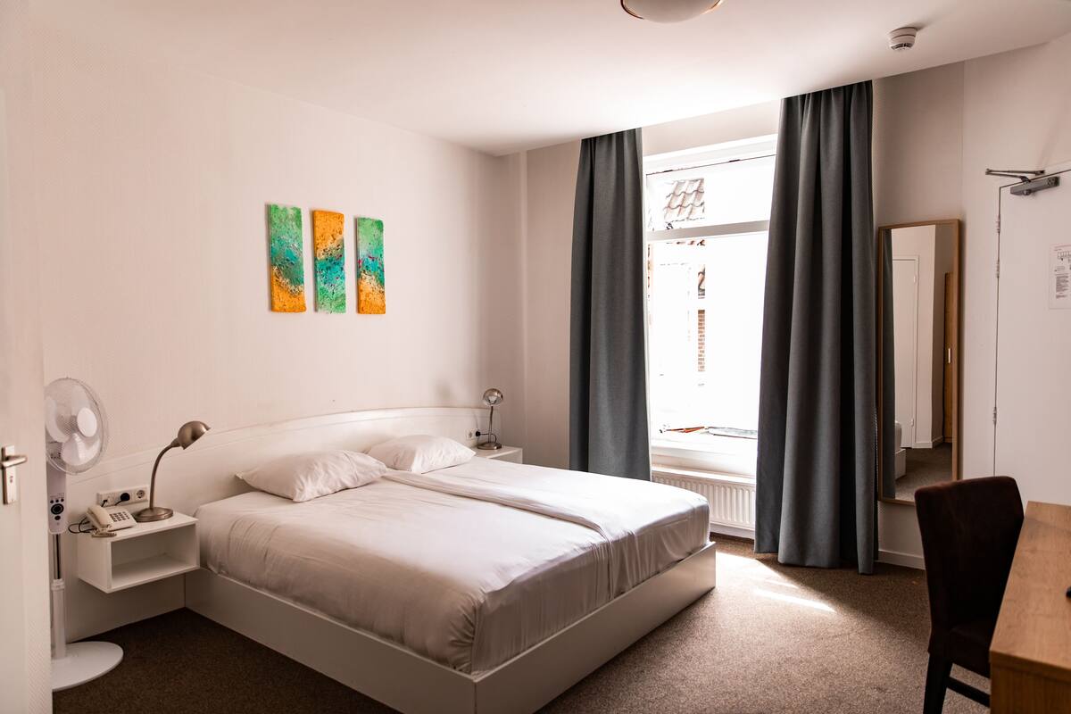 A modern hotel room with a large bed, bedside tables, a desk, and a window with curtains.