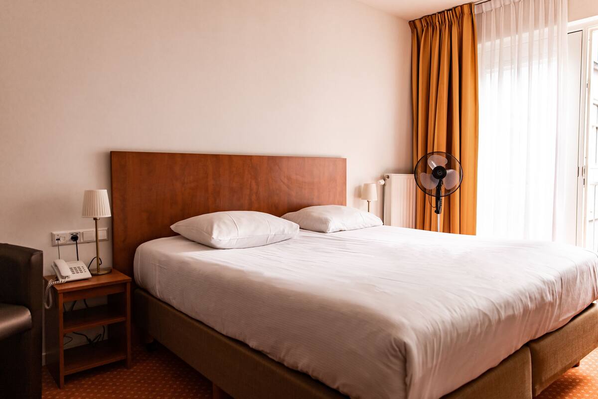 A hotel room with a wooden headboard, a bed with white linens, a bedside table with a lamp and a phone, and a standing fan.