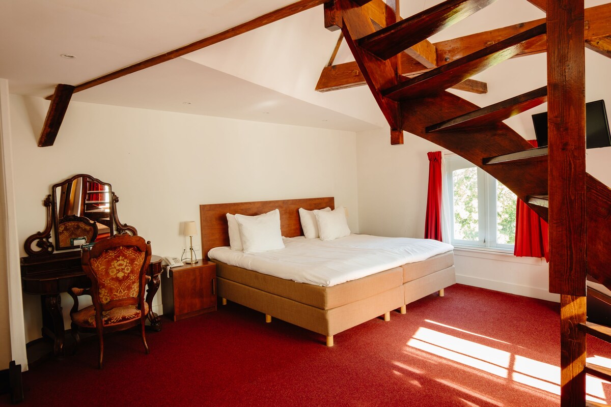 A bedroom with a wooden ceiling, a large bed, a chair, a desk, and a window with curtains.