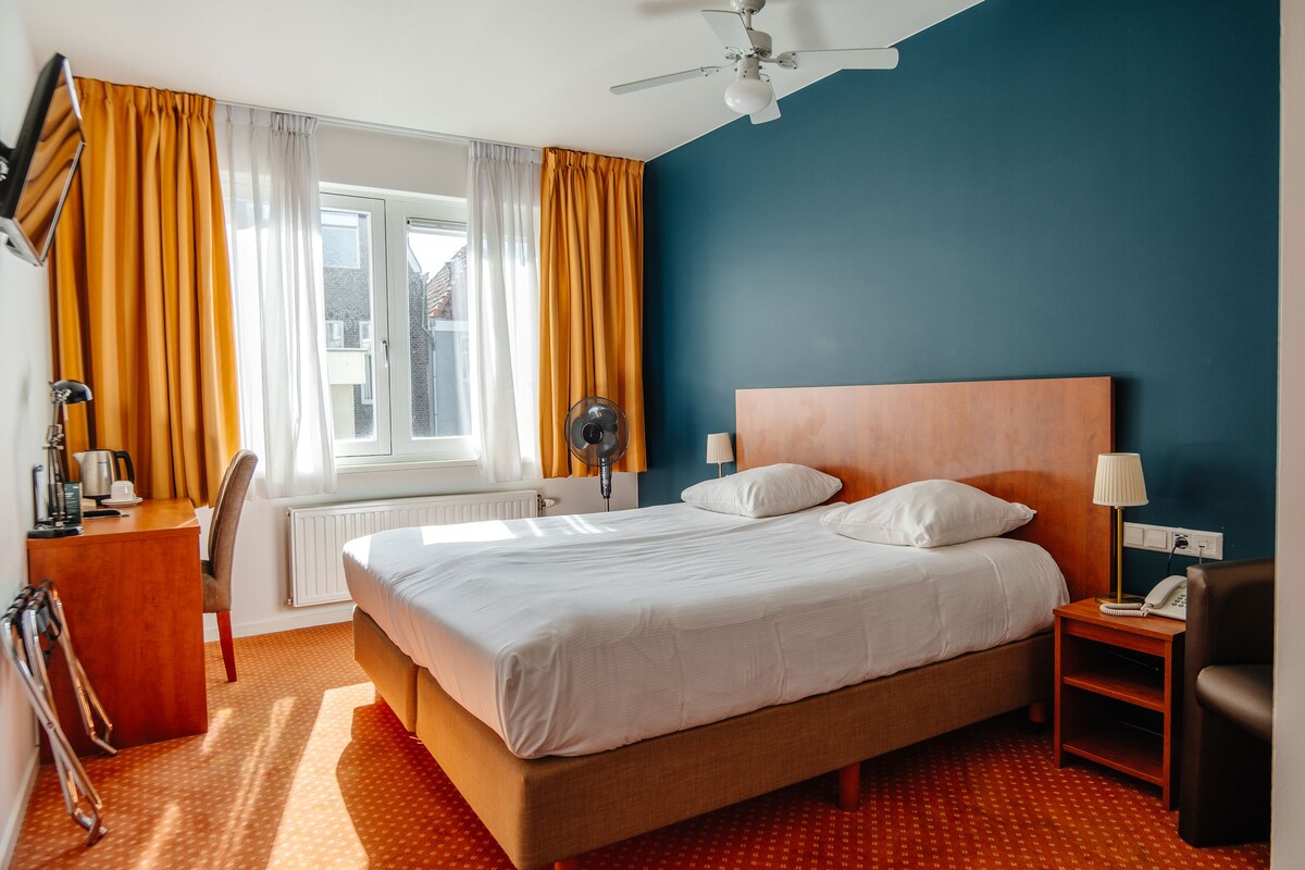 A hotel room with a bed, a desk, a chair, a nightstand, and a window with curtains.
