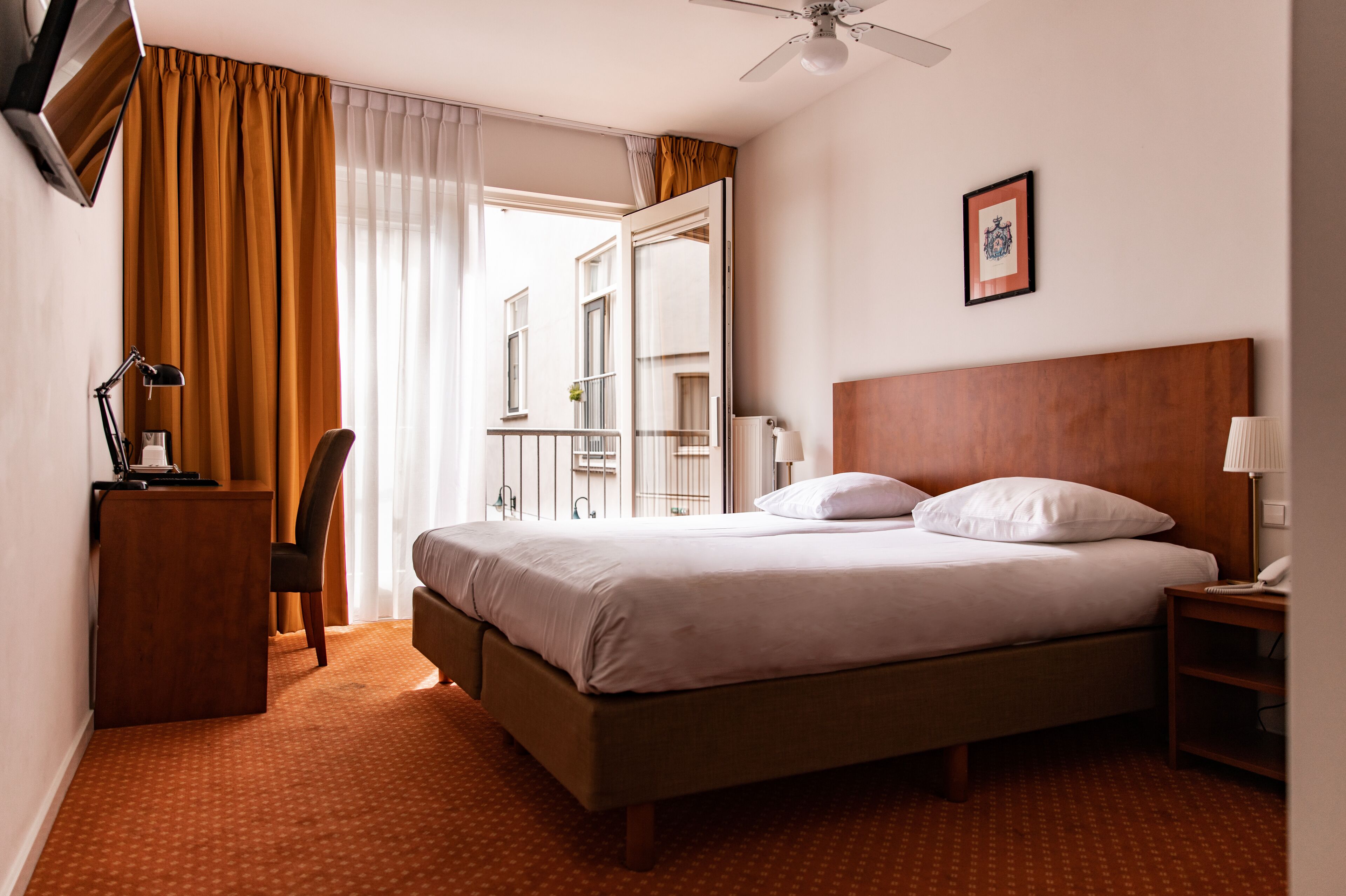 A hotel room with a bed, a desk, a chair, a nightstand, a ceiling fan, and a window with curtains.