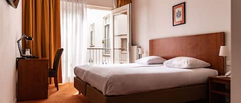 A hotel room with a bed, a desk, a chair, a nightstand, a ceiling fan, and a window with curtains.