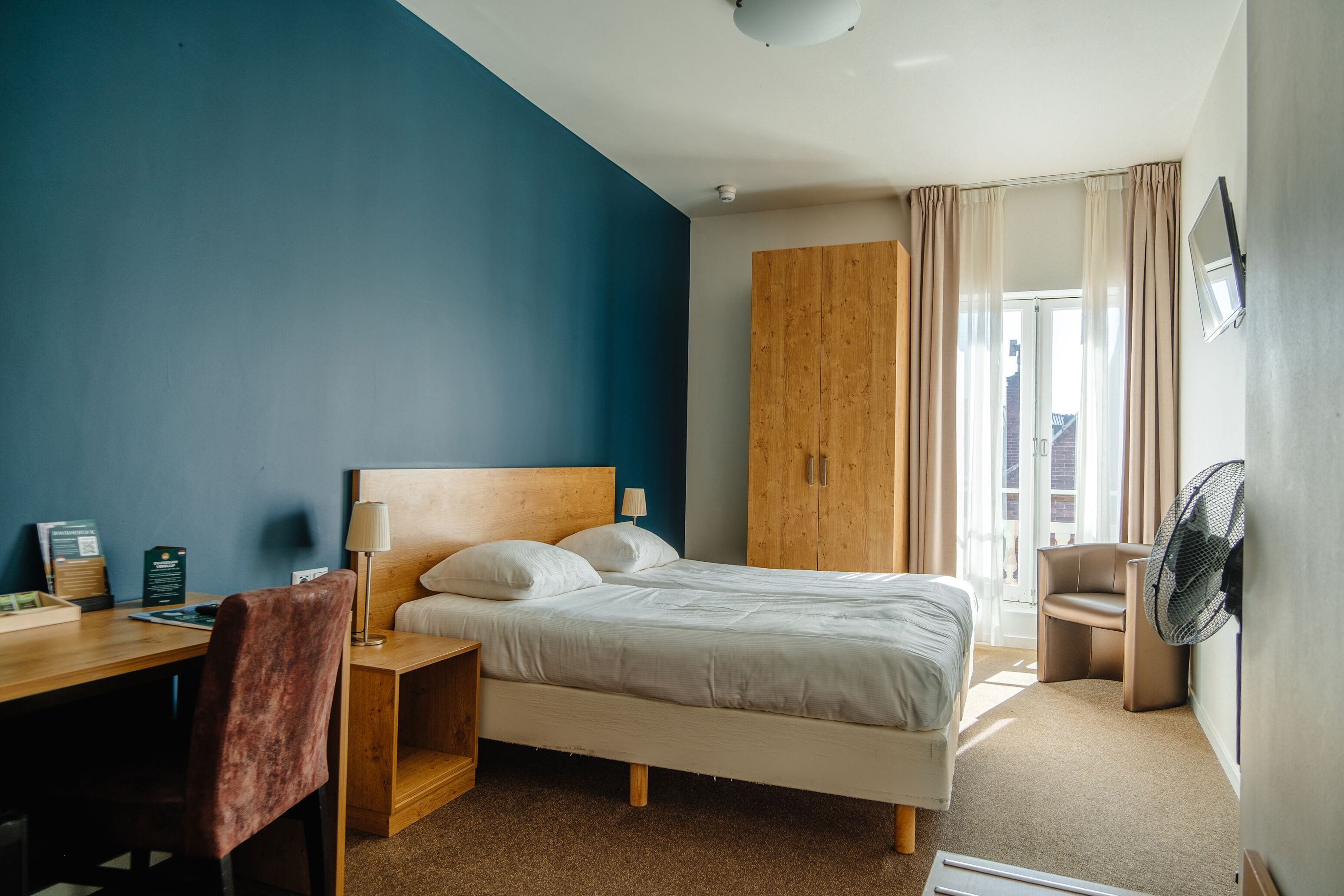 Ein Hotelzimmer mit Bett, Schreibtisch, Stuhl, Kleiderschrank und Ventilator.