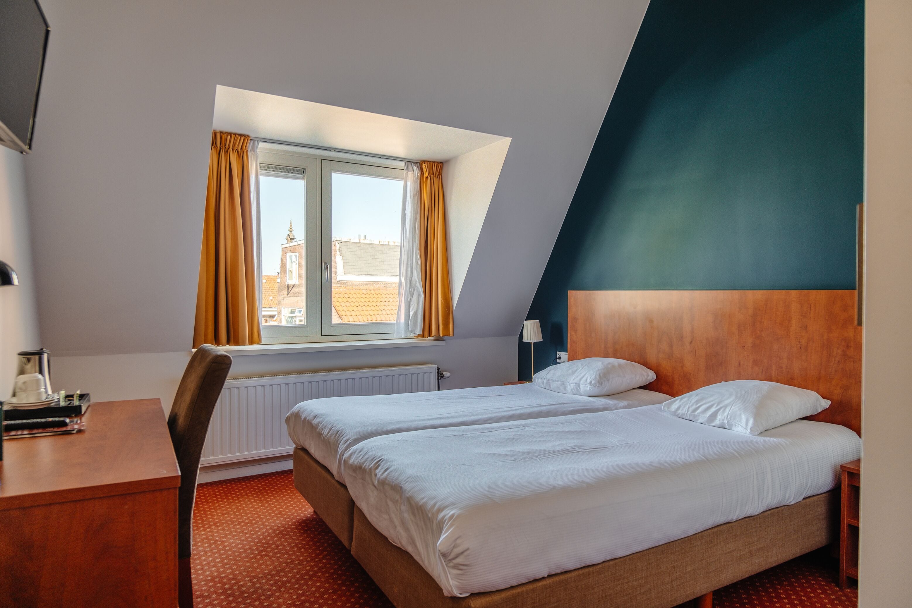 A hotel room with two beds, a desk, a chair, a window with curtains, and a view of a building outside.