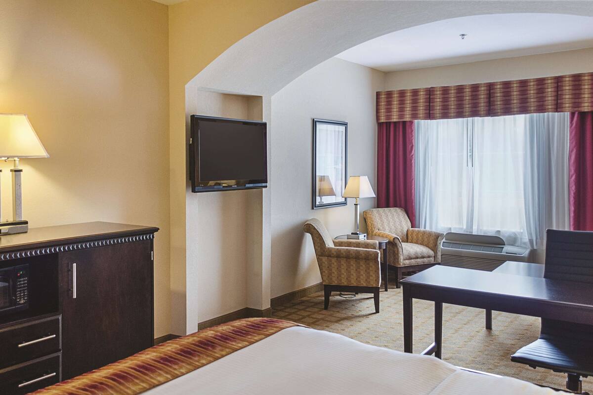 Une chambre d’hôtel comprenant un lit, deux fauteuils, un bureau, un téléviseur à écran plat, une lampe et une fenêtre avec des rideaux.