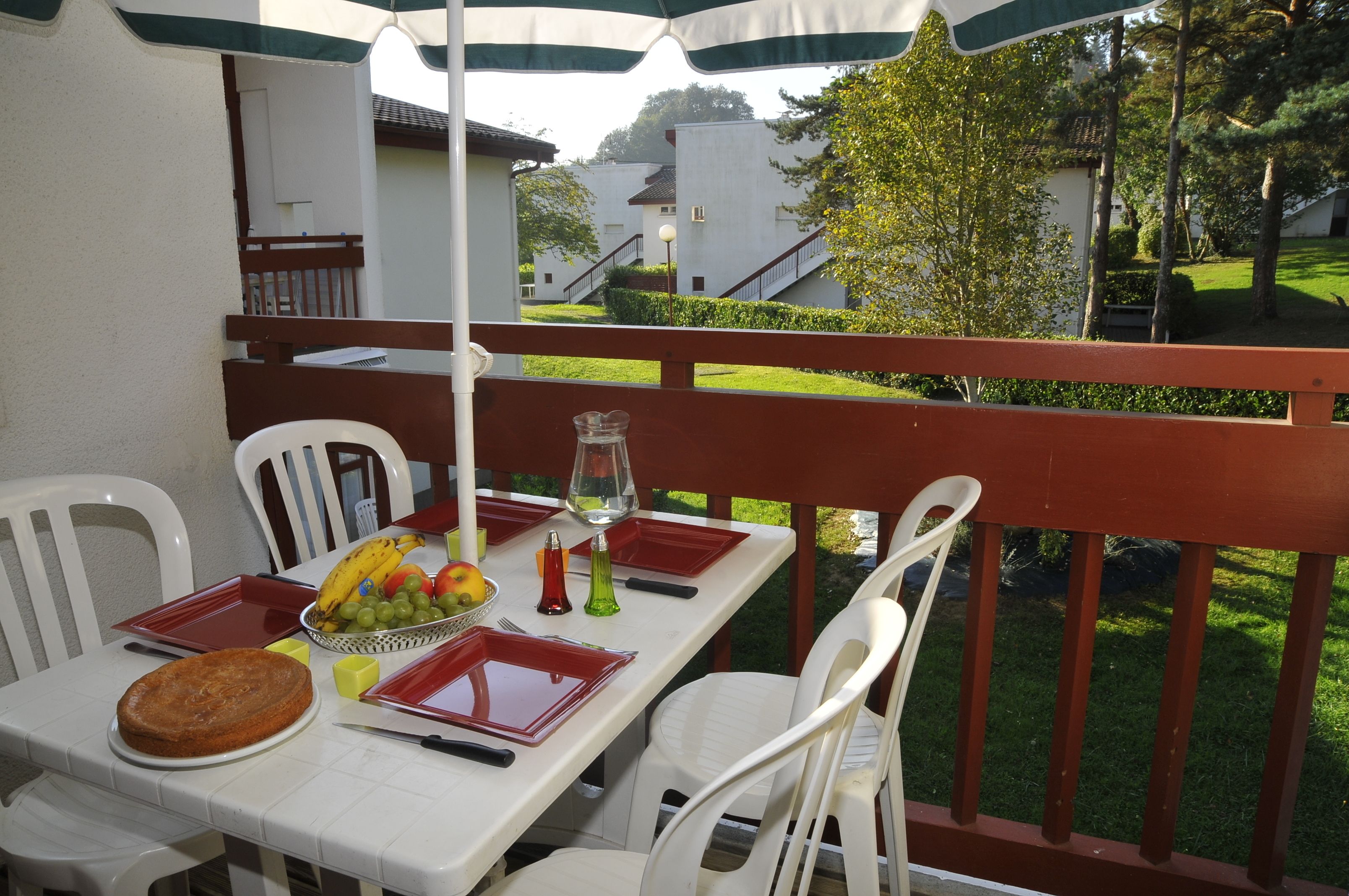 Un área de comedor al aire libre con una mesa dispuesta para dos personas, que incluye un frutero, un pastel y una copa de vino.