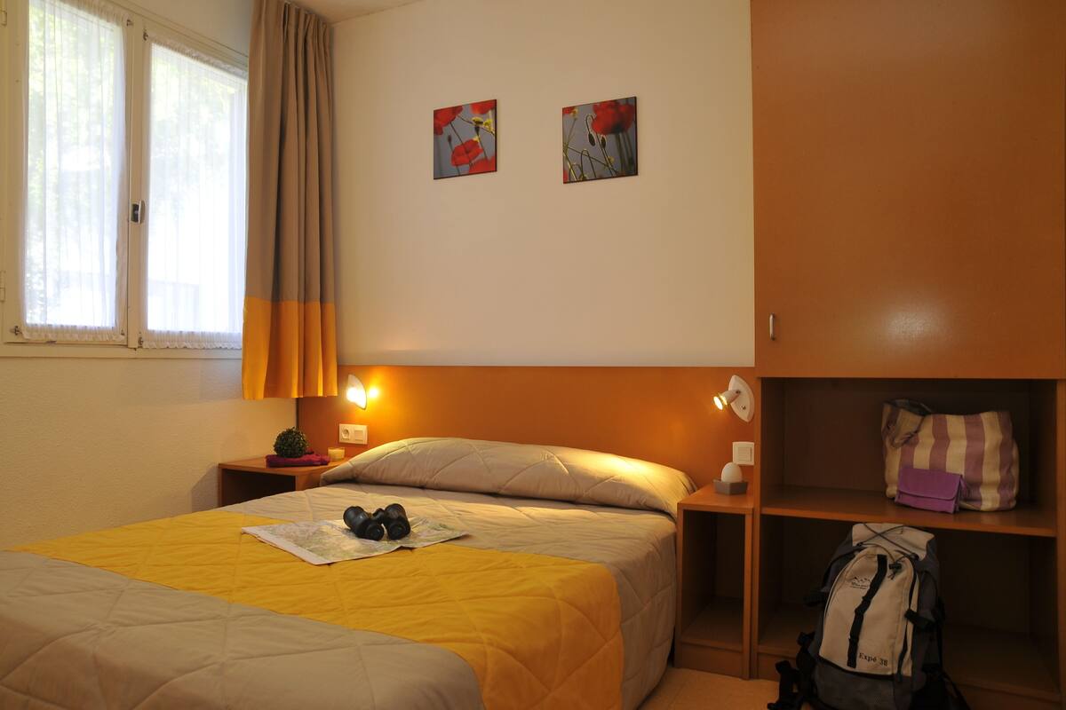 A hotel room with a bed, a nightstand, a lamp, a shelf with a bag, and two paintings on the wall.