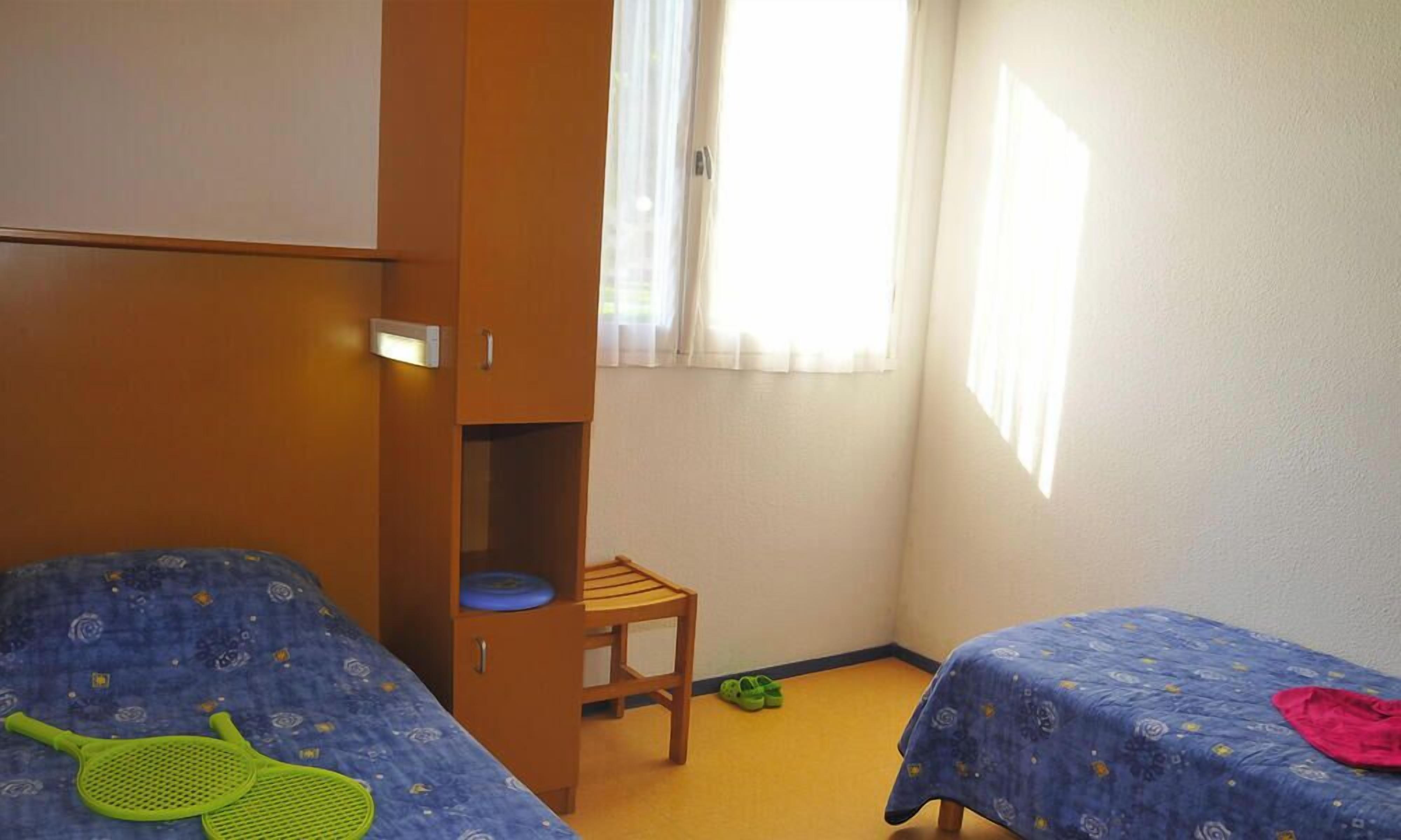 A bedroom with two beds, a wardrobe, a chair, and a window.