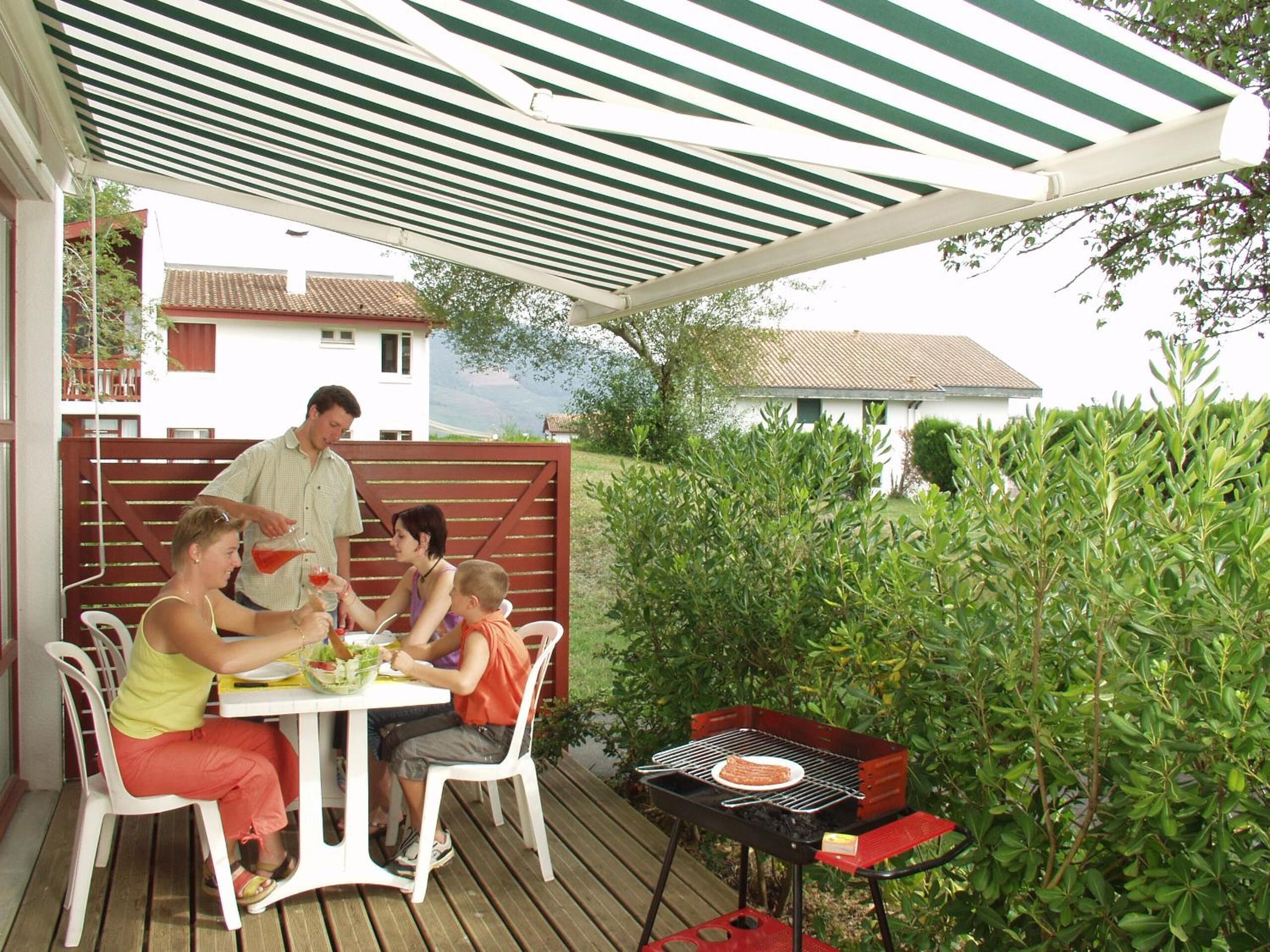 A family dining outdoors under a striped canopy, with a barbecue grill and a table set for a meal.