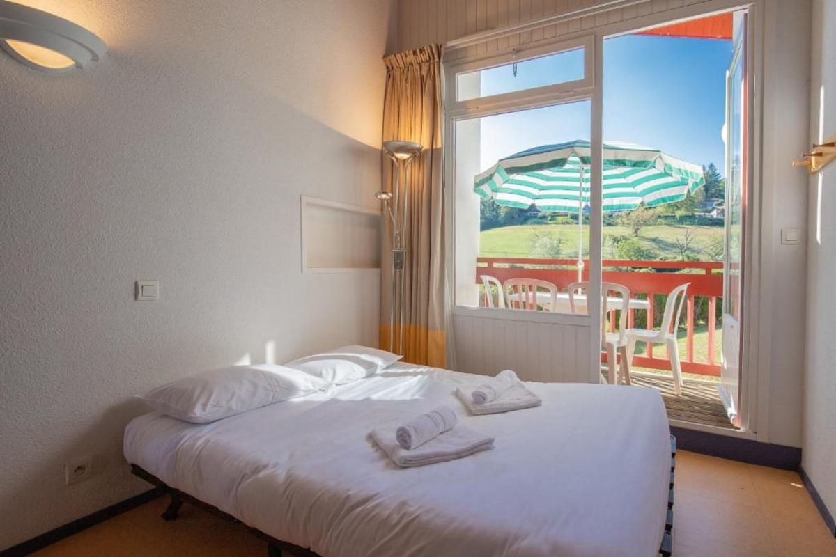 A hotel room with a bed, a window with curtains, and a view of an outdoor area with a chair and a table.