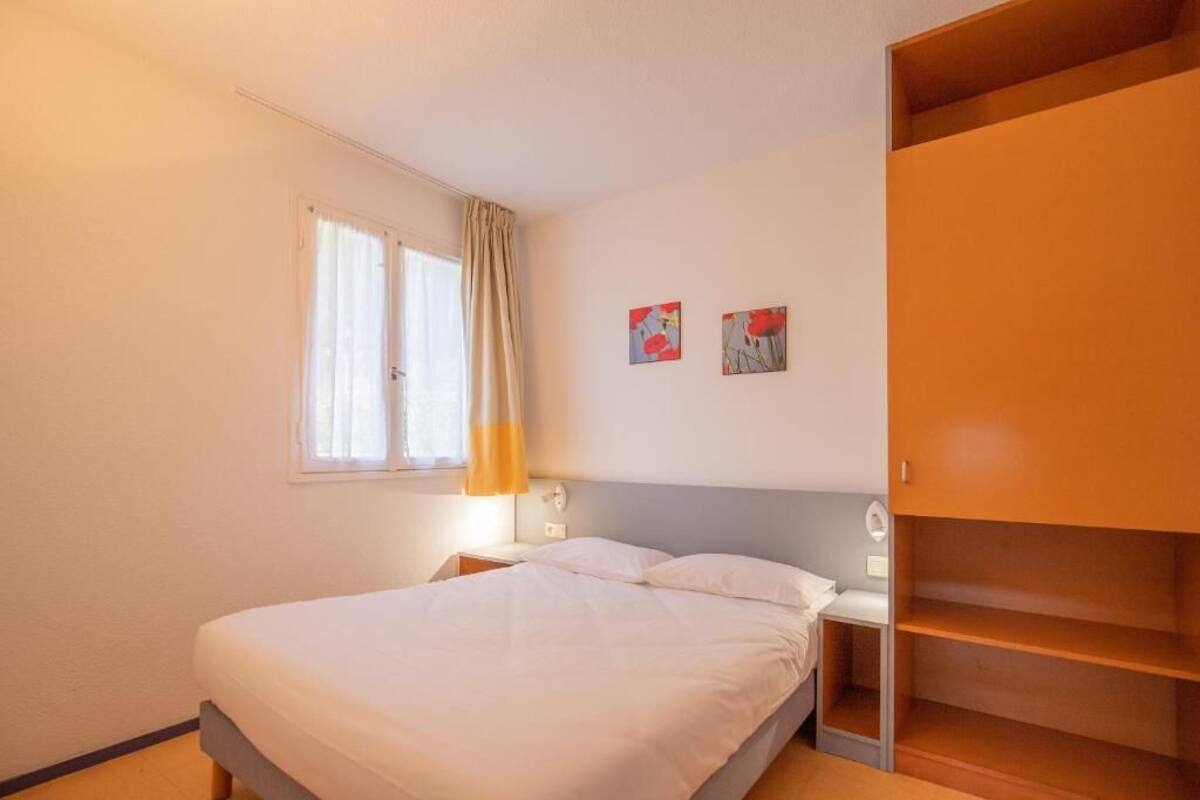 A bedroom with a bed, a nightstand, a bedside table, a window with curtains, and a wardrobe.