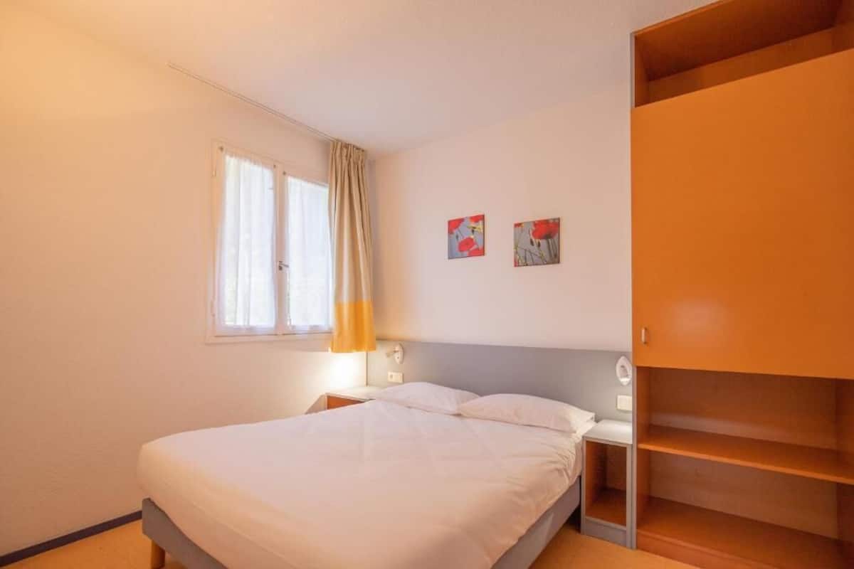 Ein Schlafzimmer mit Bett, Nachttisch, Fenster mit Vorhängen und Kleiderschrank.