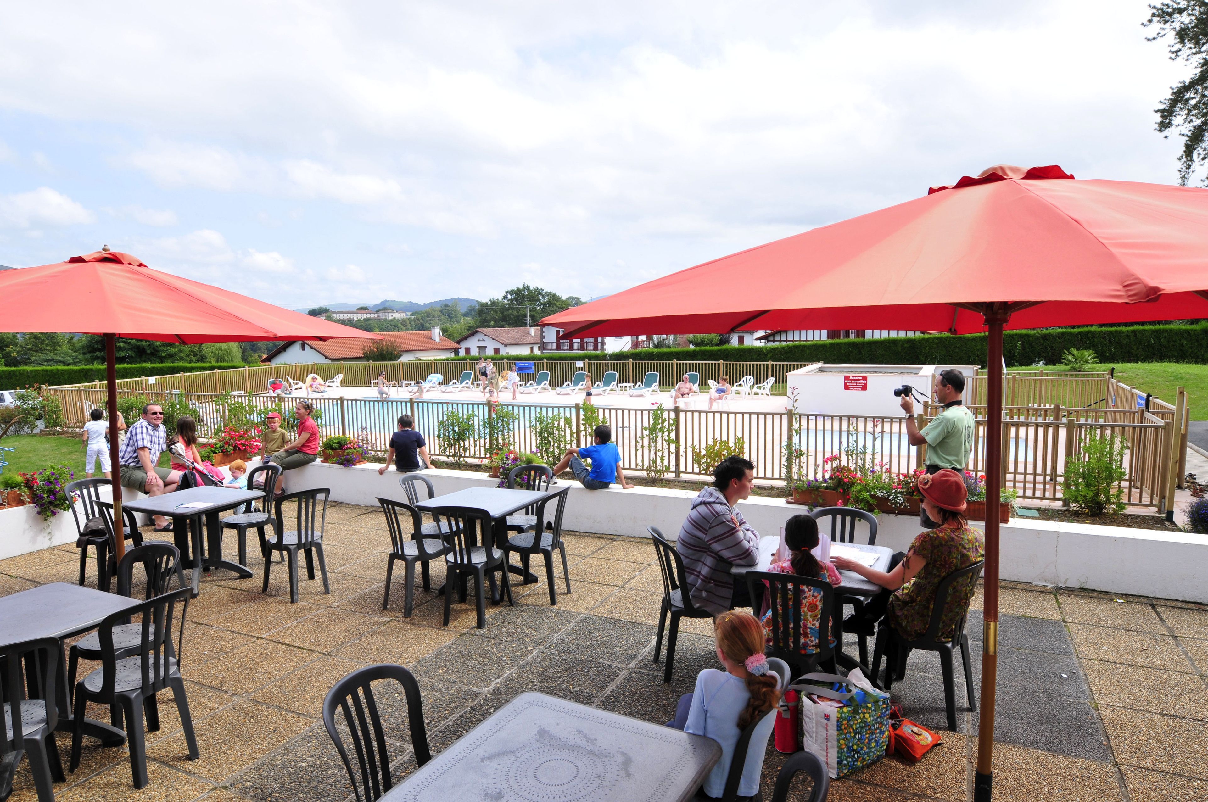 Un espace extérieur aménagé en terrasse, avec des tables et des chaises sous des parasols rouges, une piscine en arrière-plan et un jardin clos.