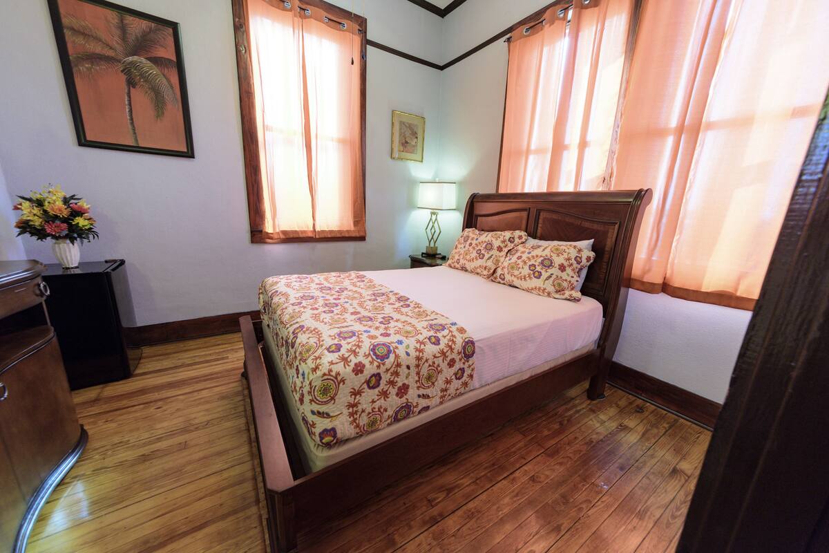 A bedroom with a wooden bed, a nightstand, a lamp, a framed picture on the wall, and a vase with flowers on a dresser.