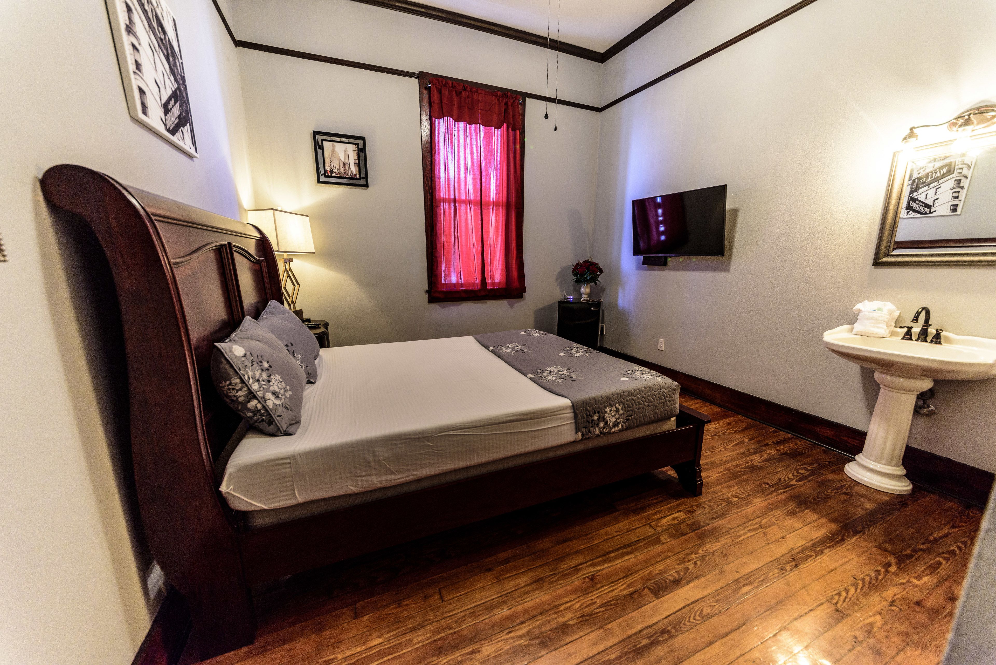 A bedroom with a wooden bed, a bedside table with a lamp, a window with red curtains, a wall-mounted TV, and a white pedestal sink.