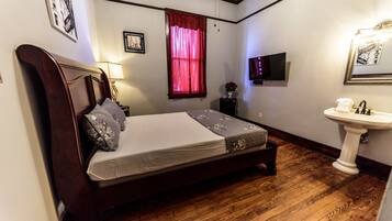A bedroom with a wooden bed, a bedside table with a lamp, a window with red curtains, a wall-mounted TV, and a white pedestal sink.