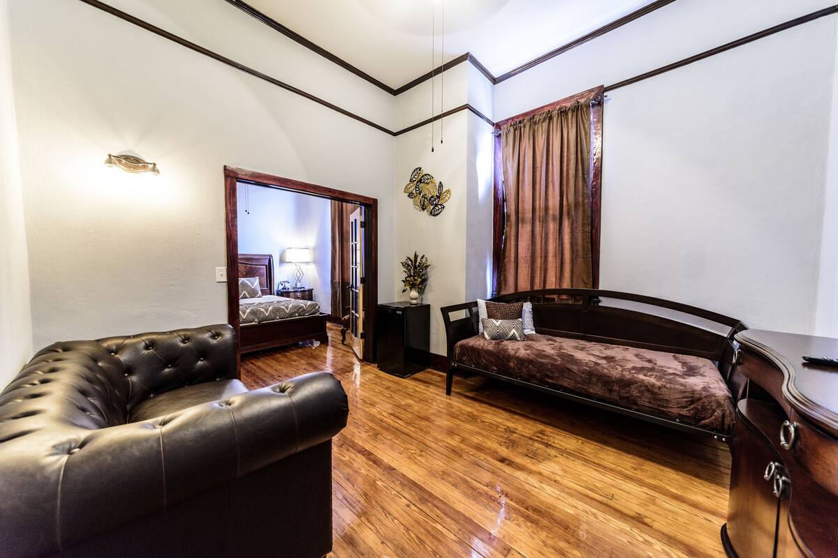 A room with a leather sofa, a bed with a headboard, a small table, and a wall-mounted lamp.
