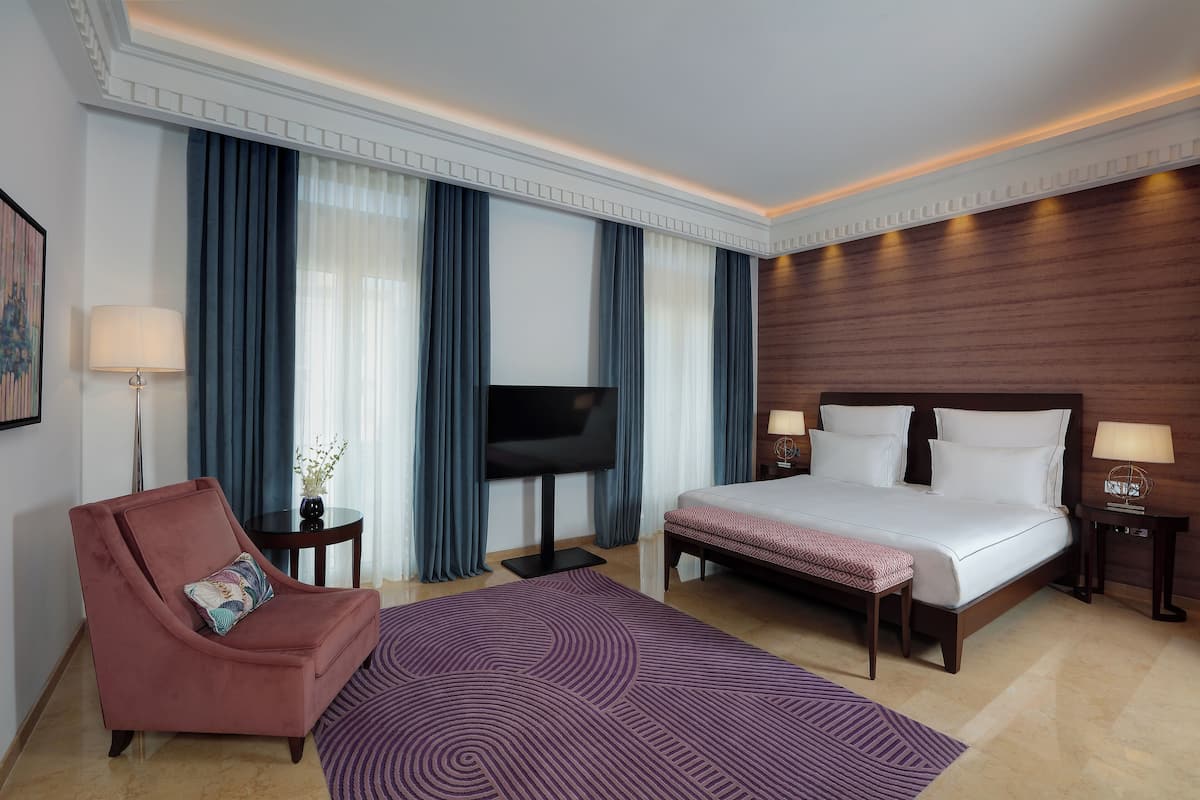 Habitación de hotel con cama, sillón, televisión y escritorio con lámparas.