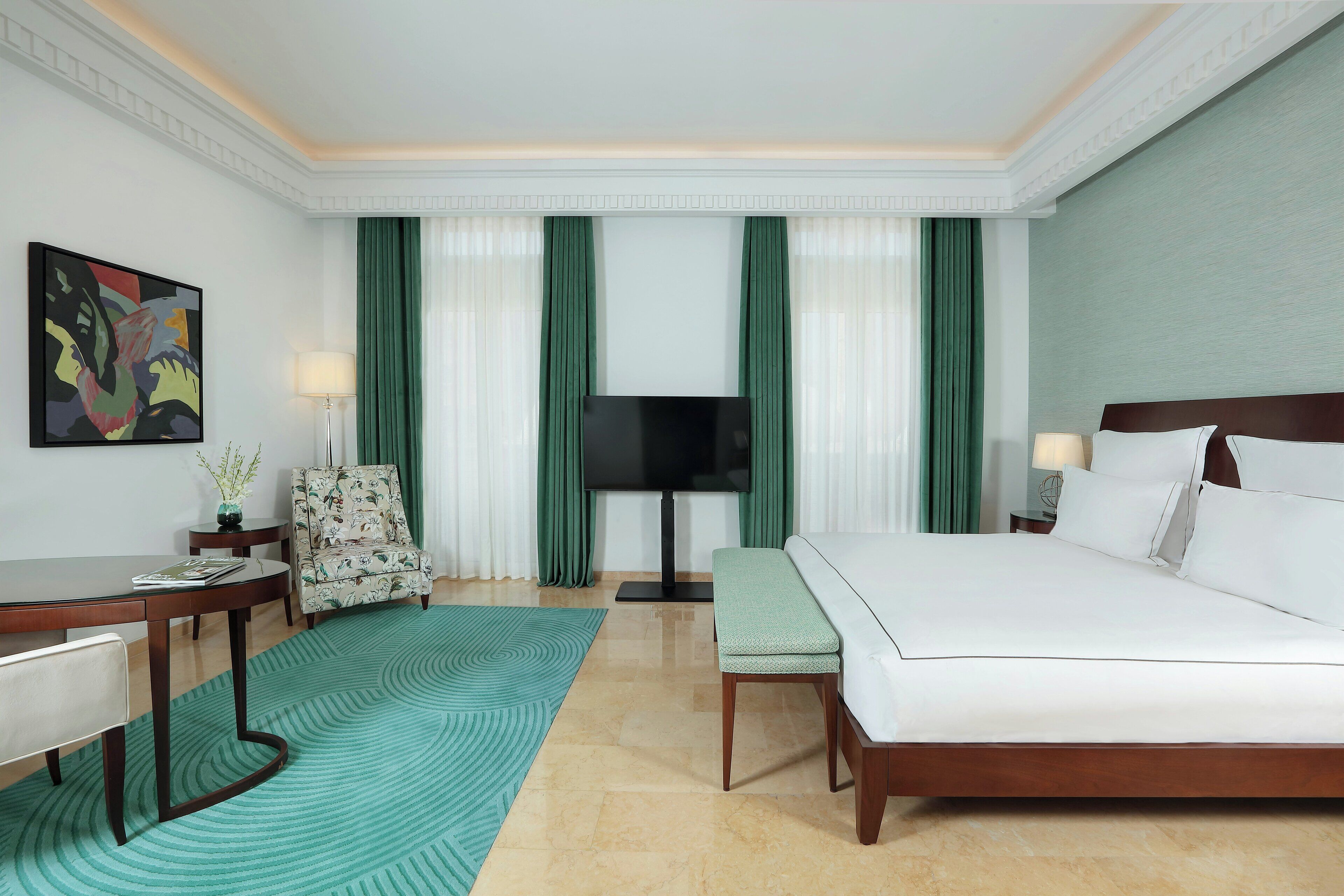 A hotel room with a bed, a desk, a chair, a TV, and a painting on the wall.