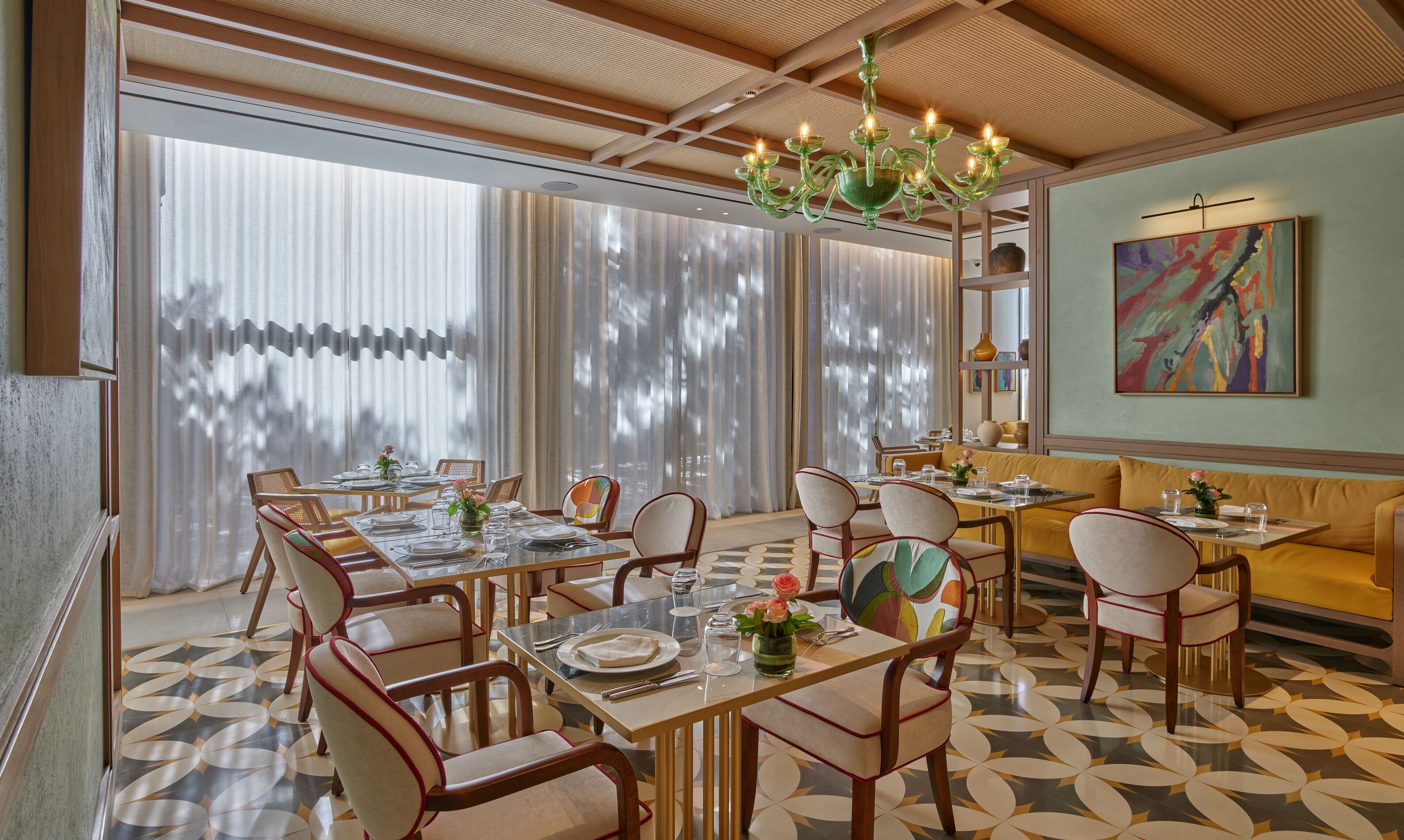A dining area with tables set for four, wooden chairs, a patterned floor, and a large window with sheer curtains.