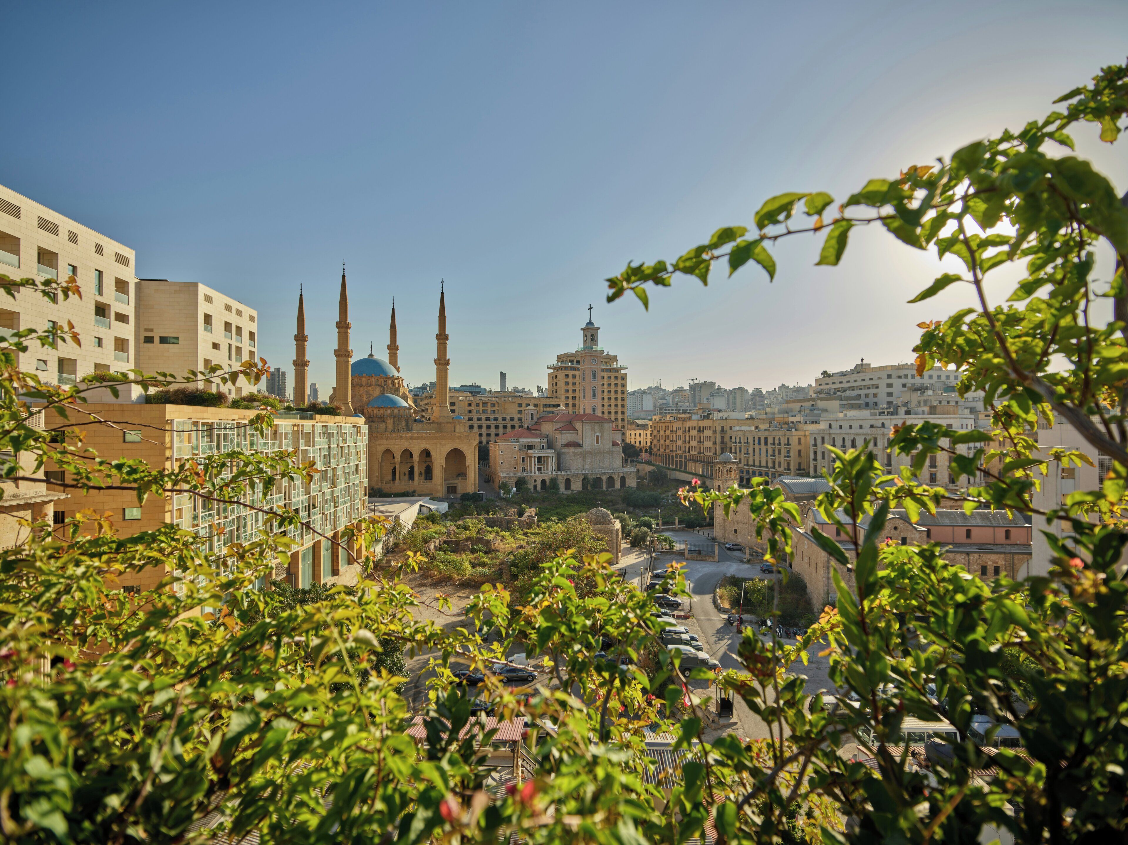 A cityscape with a prominent mosque with multiple minarets and domes, surrounded by modern buildings and greenery.
