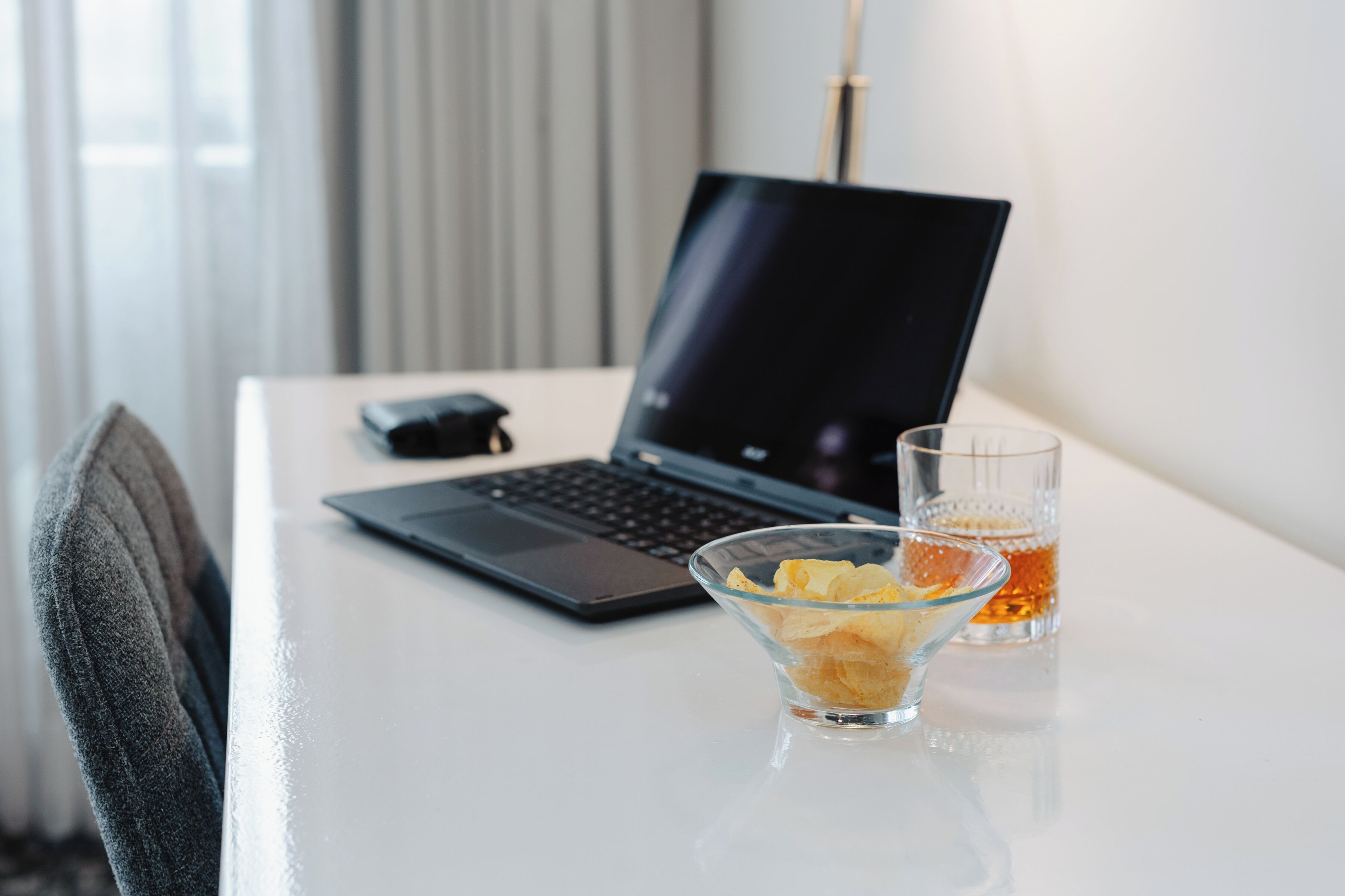 Una laptop sobre un escritorio blanco, con un vaso de líquido ámbar y un tazón de bocadillos.