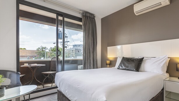 Une chambre d’hôtel avec un grand lit, une porte vitrée donnant sur un balcon, une petite table avec une chaise et une table de chevet avec une lampe.