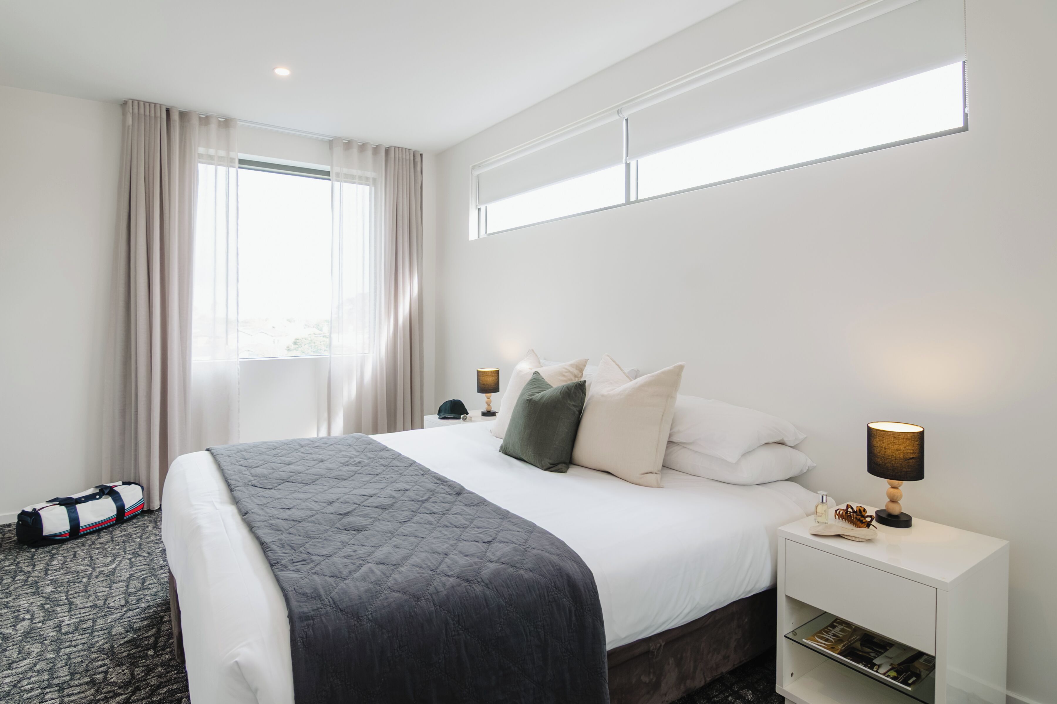 A hotel room with a bed, two bedside lamps, a nightstand, a window with curtains, and a bag on the floor.