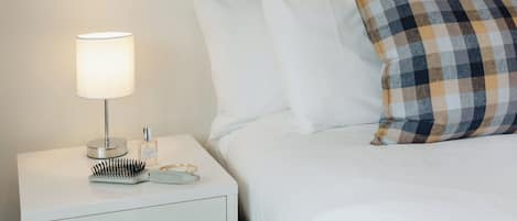 A neatly made bed with white linens, a plaid pillow, a white bedside table with a lamp, and a hairbrush.