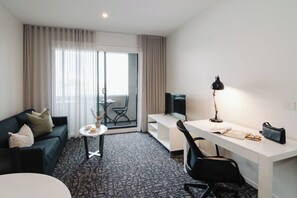 A hotel room with a sofa, a small table, a desk with a chair, a TV, and a balcony with curtains. - Punthill Oakleigh (Hughesdale)