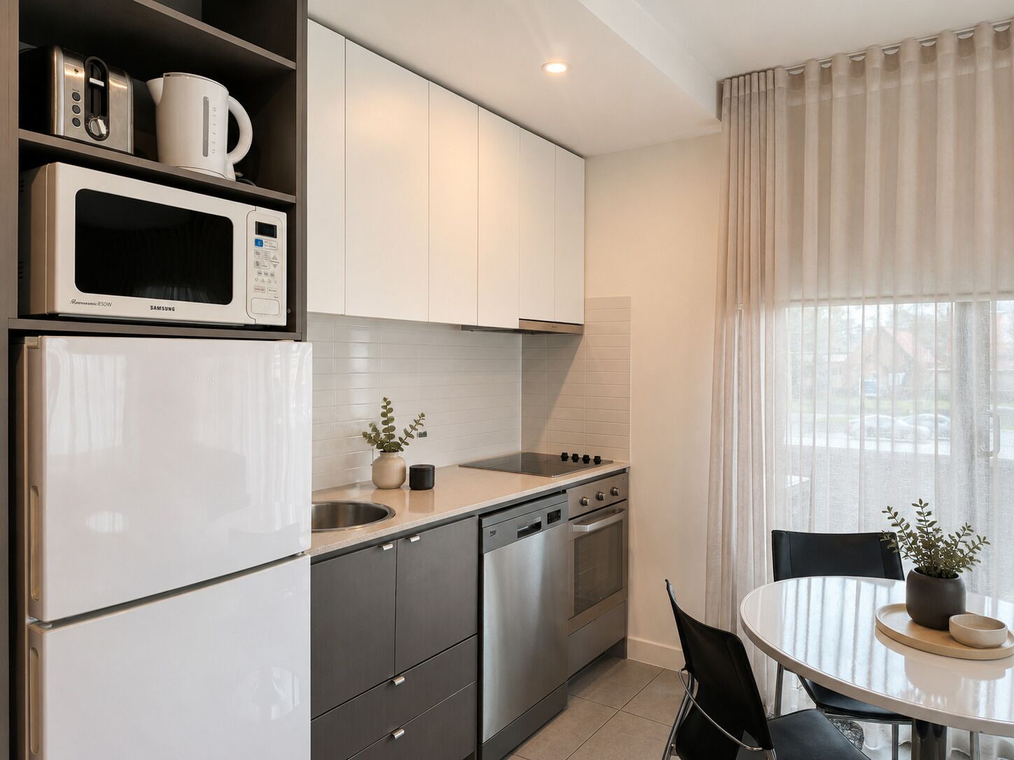 Una cocina moderna con armarios blancos, electrodomésticos de acero inoxidable, una zona de comedor con mesa y sillas, y una ventana con cortinas.