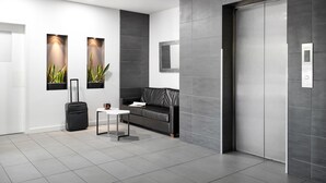 Un hall d’entrée moderne avec un sol carrelé de couleur grise, un canapé en cuir noir, une petite table, une valise noire et une plante fixée au mur.