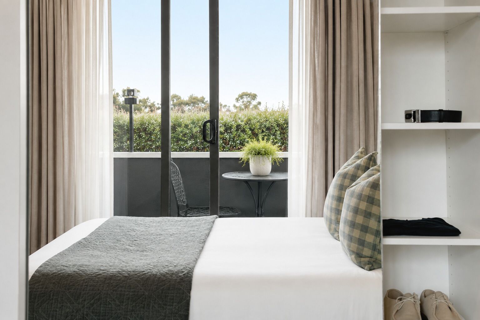 Habitación de hotel con cama, un pequeño balcón con mesa y silla, una planta en maceta y un zapatero.