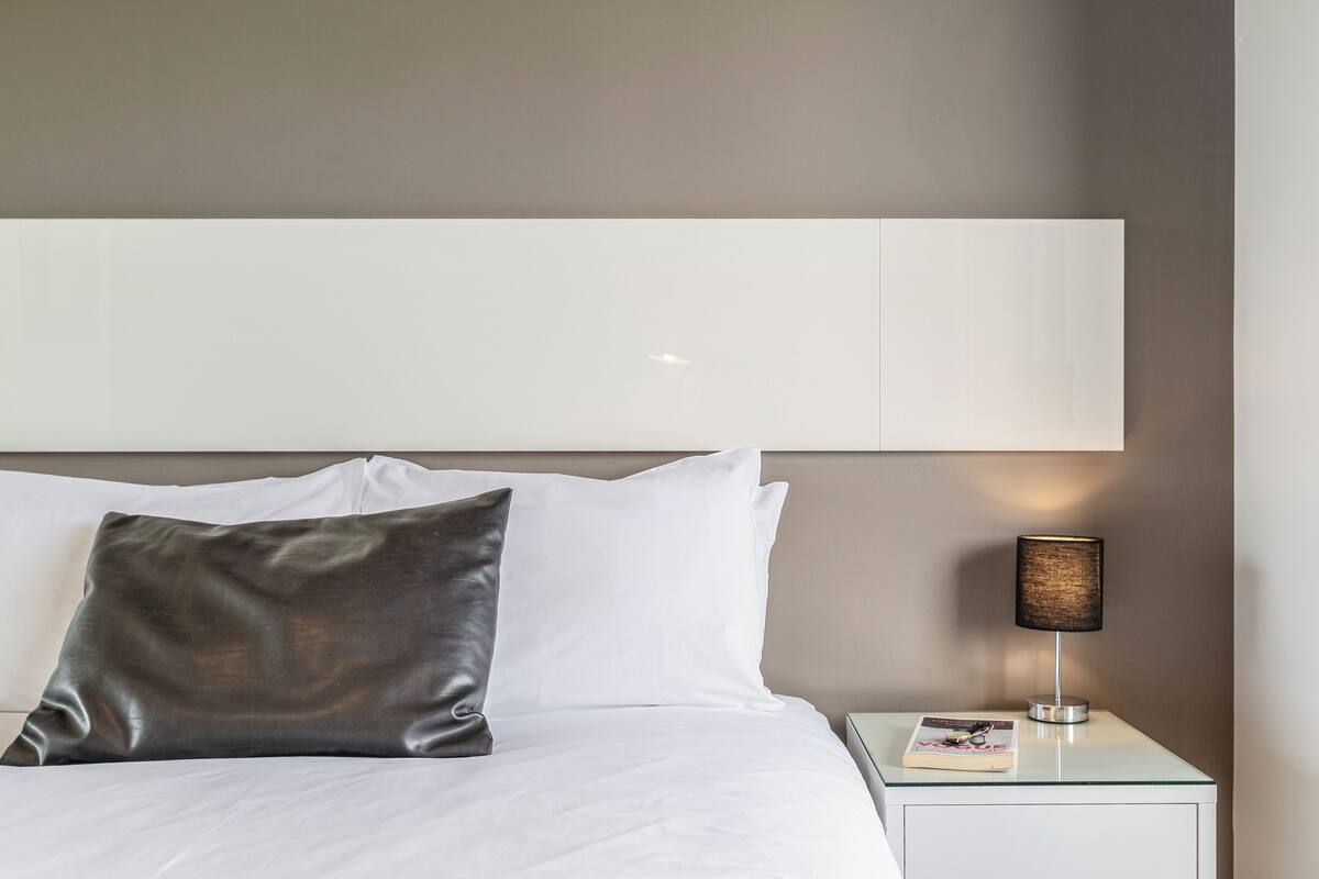 A neatly made bed with white linens and a single dark pillow, a bedside lamp, and a book on a small bedside table.