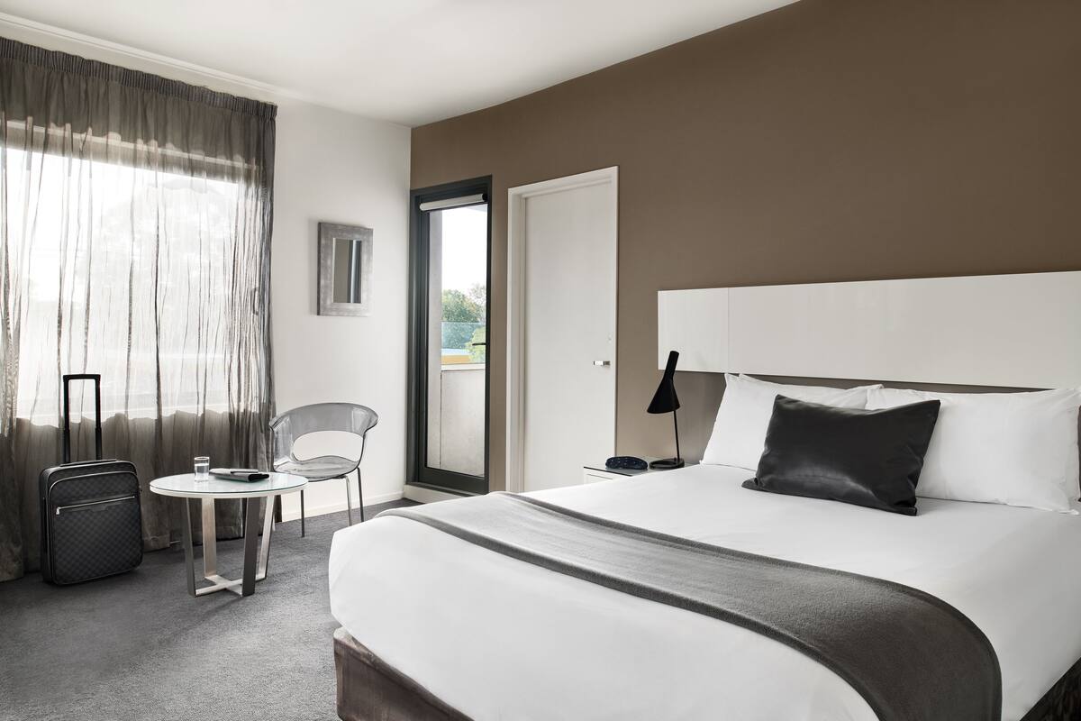A hotel room with a large bed, a small table with a chair, a suitcase, and a door leading to a balcony.