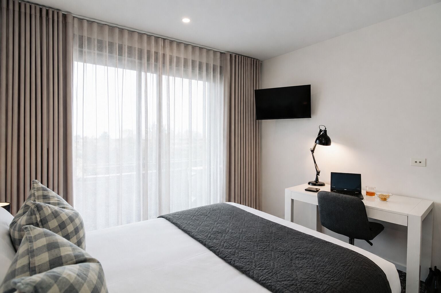 A hotel room with a bed, a desk with a computer, a TV, and a window with curtains.